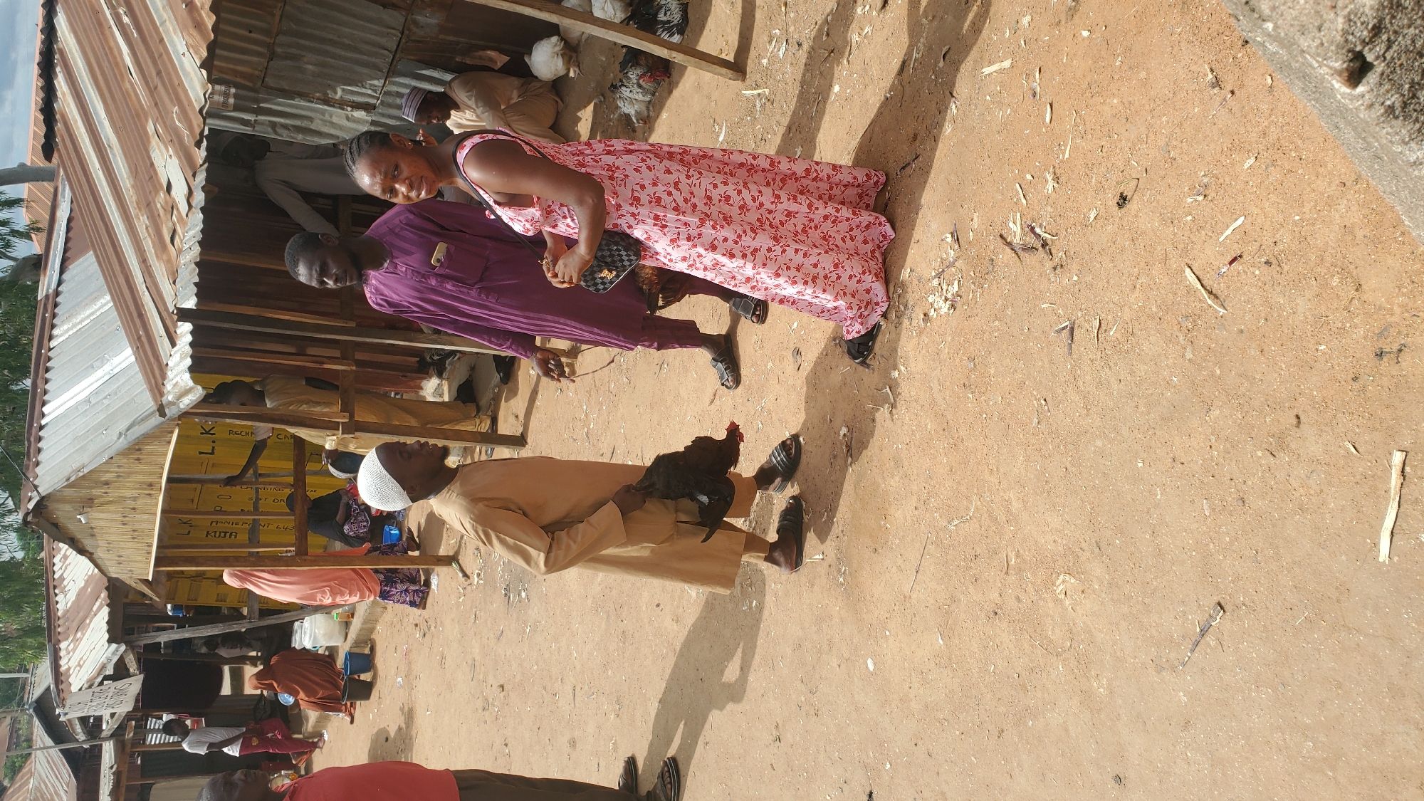 The image depicts an outdoor market scene in Jalingo, Nigeria. In the foreground, a young boy wearing a white cap and a tan robe is holding a dead black chicken by its legs. To his right, a woman in a pink floral dress is looking towards the camera and holding a black handbag. Behind them, a man in a purple robe stands looking to the left. The background shows several small market stalls with corrugated metal roofs. Several other people and chickens are visible further back within the market. The ground is dirt and scattered with debris.