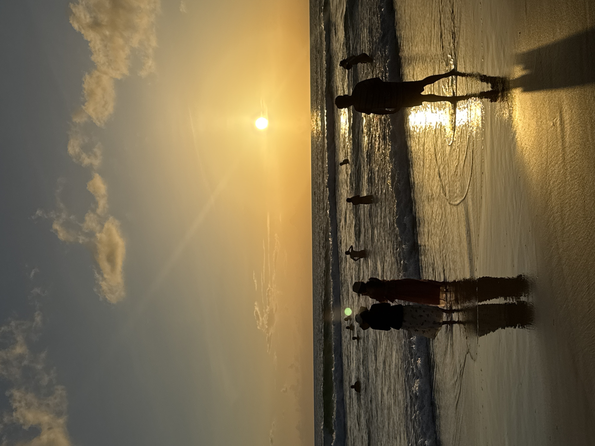 The image depicts a sunset scene on Karon Beach in Karon, Thailand. Silhouetted figures are visible on the wet sand and in the shallow ocean water. In the foreground, two individuals stand facing the ocean, their backs to the camera, with their reflections cast on the wet sand. To their right, a solitary figure stands in the water, also silhouetted against the sunset. Further out in the ocean, several other individuals are wading or swimming. The sun, a bright orb, is setting on the horizon, casting a warm golden glow over the water and sky. The ocean is calm with gentle waves lapping at the shore, and a few scattered clouds dot the sky. The wet sand in the foreground is smooth and reflective, mirroring the light from the sunset and the figures.