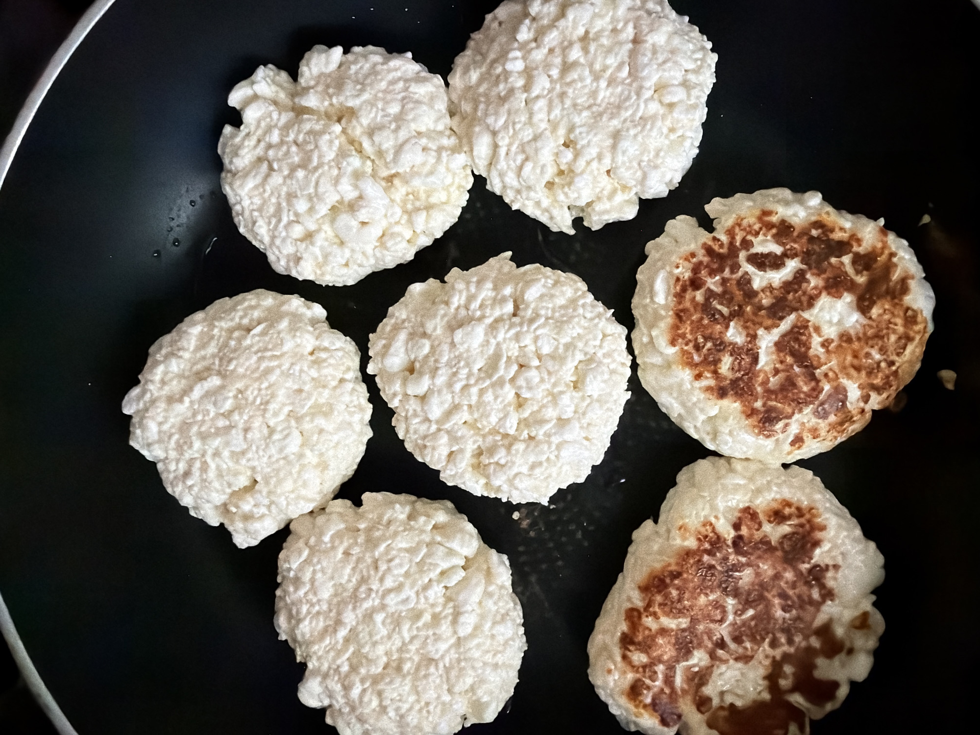 A close-up, top-down view of seven uncooked cheese pancakes being fried in a black non-stick frying pan. Five of the pancakes are pale white and uncooked, while the bottom two have begun to brown on one side. The frying pan is clean except for a few small water droplets. This image was taken in Khabarovsk, Russia.