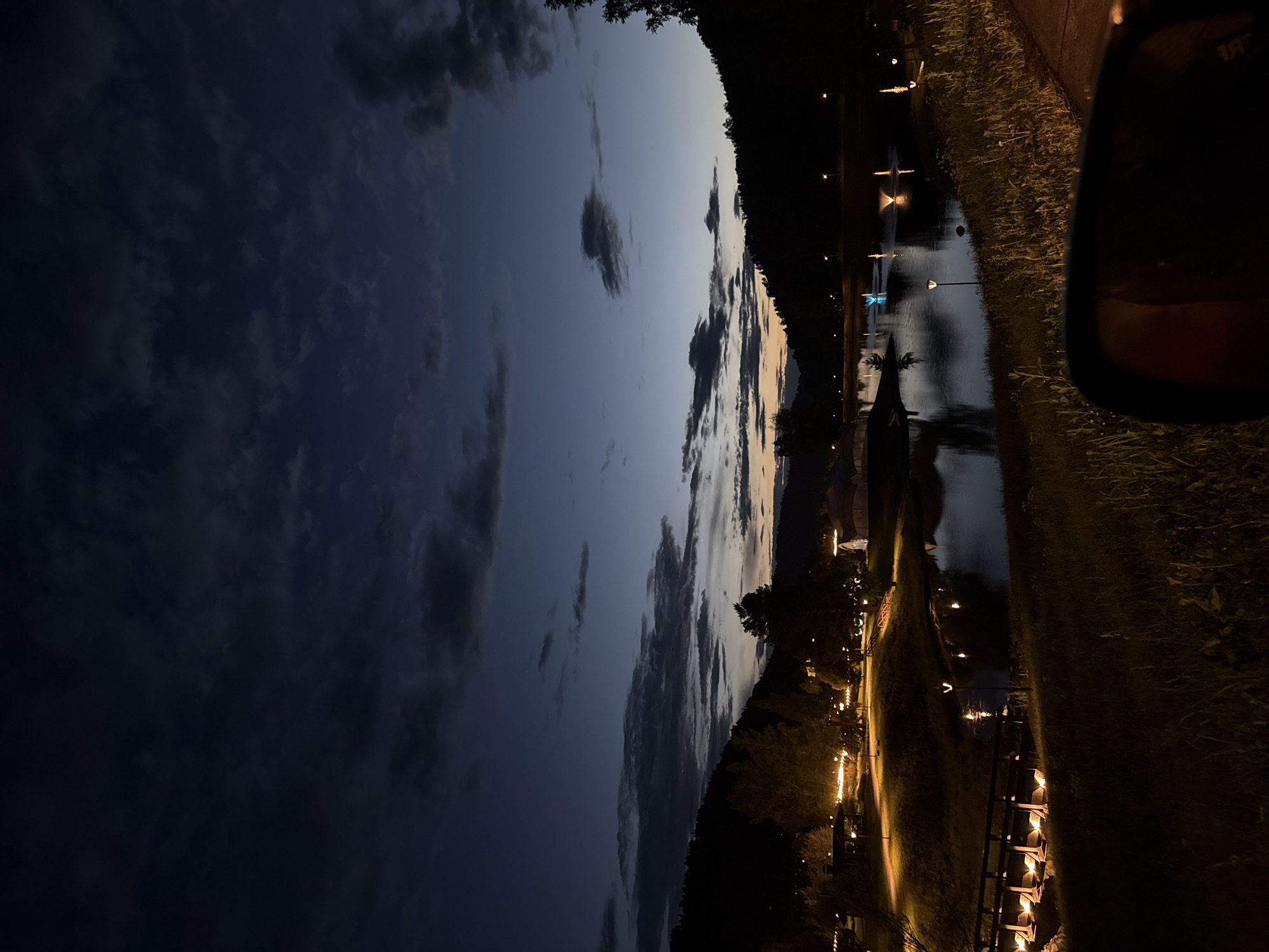 The image depicts a twilight scene at a resort or park area, likely in the city of Urlu-Aspak, Russia. The sky is a deep blue, transitioning to a pale orange and yellow hue near the horizon, indicating the end of sunset. Dark clouds are scattered across the sky. In the foreground, a grassy embankment slopes down towards a body of water. To the left, a wooden bridge is illuminated by strings of warm lights. Further back, several trees are visible, also lit by decorative lights. Across the water, on the opposite bank, are more trees and the silhouettes of buildings, including what appears to be a white tent-like structure. The water itself reflects the lights from the shore and the sky, with several illuminated fountains creating colorful displays in the middle of the lake. A car's side mirror is partially visible in the bottom right corner, suggesting the photo was taken from within a vehicle. The overall atmosphere is serene and picturesque.