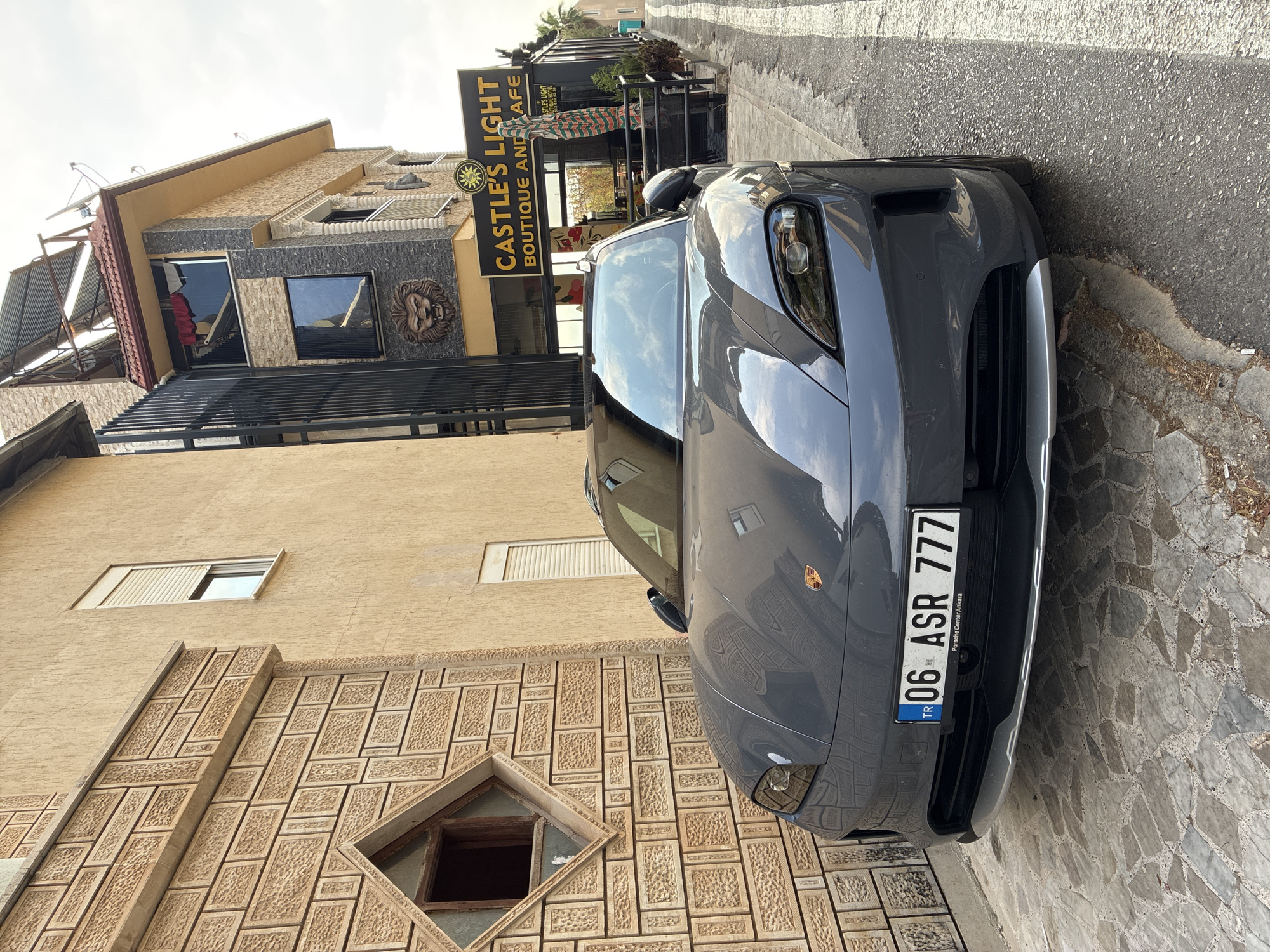 In the city of Alanya, Turkey, a dark gray Porsche Taycan is parked on a cobblestone surface in front of a building. The building features textured tan and grey stone cladding and a sign that reads "CASTLE'S LIGHT BOUTIQUE AND CAFE." The car's license plate, "06 ASR 777," is clearly visible. Sunlight reflects off the car's surface and the surrounding buildings, casting shadows. The scene appears to be a street-level view on a partly cloudy day.