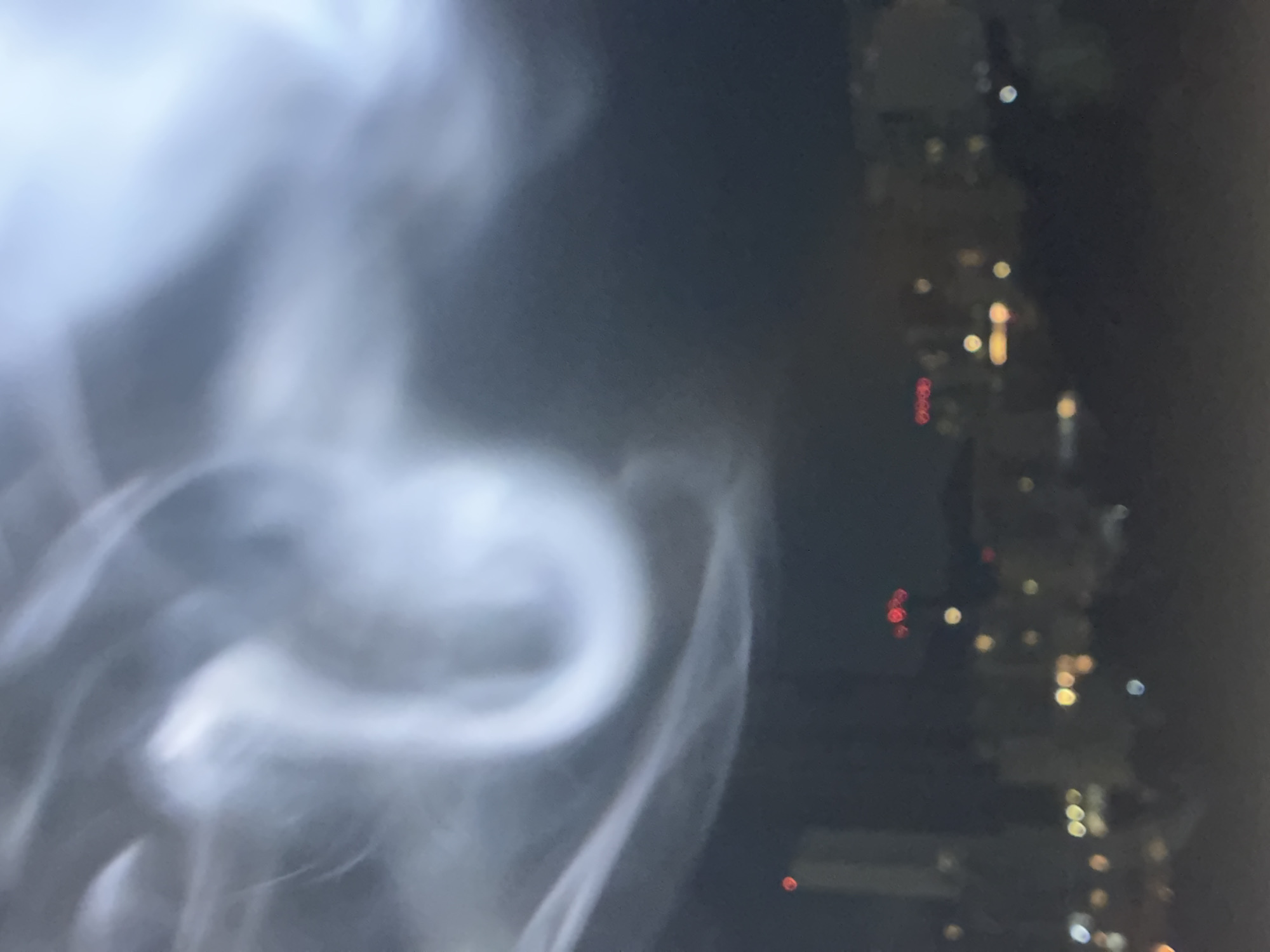 The image shows a blurry view of the city of Huixquilucan de Degollado, Mexico, at night, with smoke in the foreground. The city lights appear as blurred bokeh in the background. The smoke is dense and white, obscuring the view.