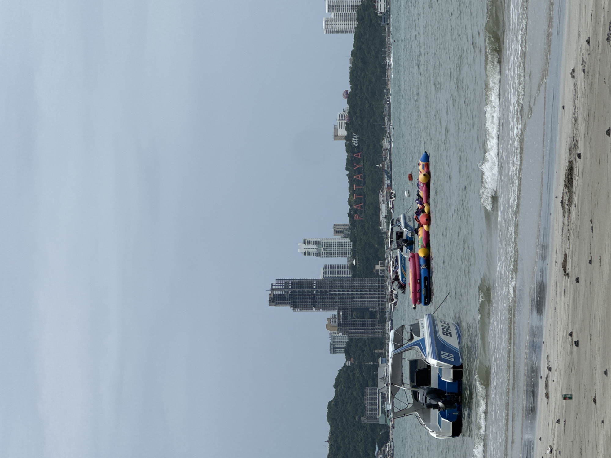 The image shows a body of water with several boats and inflatable water toys. In the background, there is a cityscape with tall buildings, including one under construction. A prominent sign on a green hillside reads "PATTAYA city". The foreground is a sandy beach with some scattered debris. The sky is overcast.