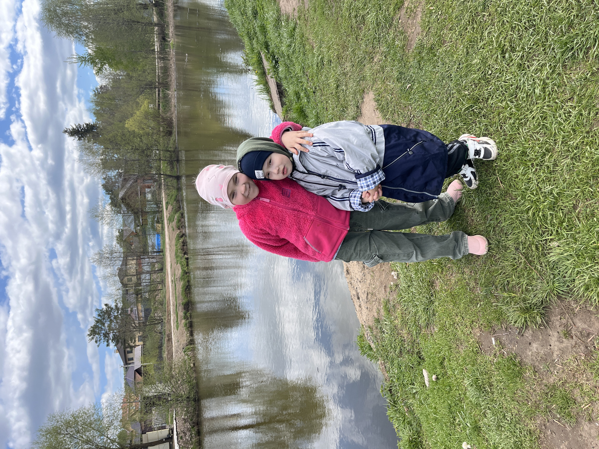 Two young children are standing on a grassy and sandy bank next to a body of water, likely a river or lake. The child on the left wears a pink hat, a pink fleece jacket, and green pants. The child on the right wears a dark blue beanie, a light gray jacket over a blue and white checkered shirt, and dark blue pants. They are in a close embrace. The body of water reflects the sky with clouds and the trees along the opposite bank. In the background are residential buildings. The location is in the city of Chaikovsky, Russia, judging from the photo context.