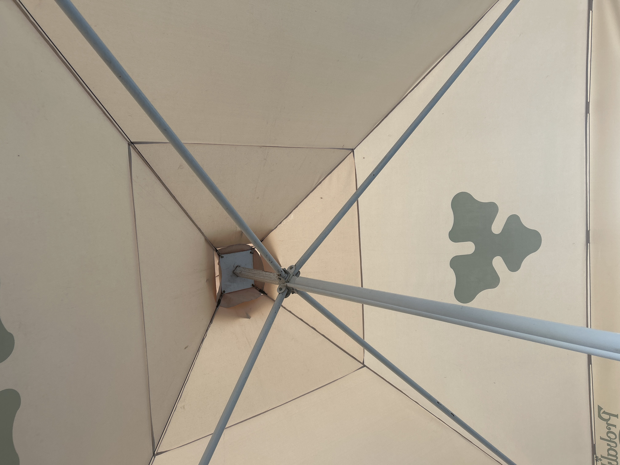 The image shows the underside of a light-colored umbrella with a symmetrical, geometric design. Several grey metal poles extend from a central hub, supporting the umbrella's fabric. The fabric is divided into triangular panels, with dark seams between them. A light green, abstract blob-like pattern is visible on one of the panels. At the bottom of the image, text is partially visible, appearing to be a logo or branding. The scene is lit with natural light, suggesting it is outdoors. There is no indication of the specific location within Brovary, Ukraine.