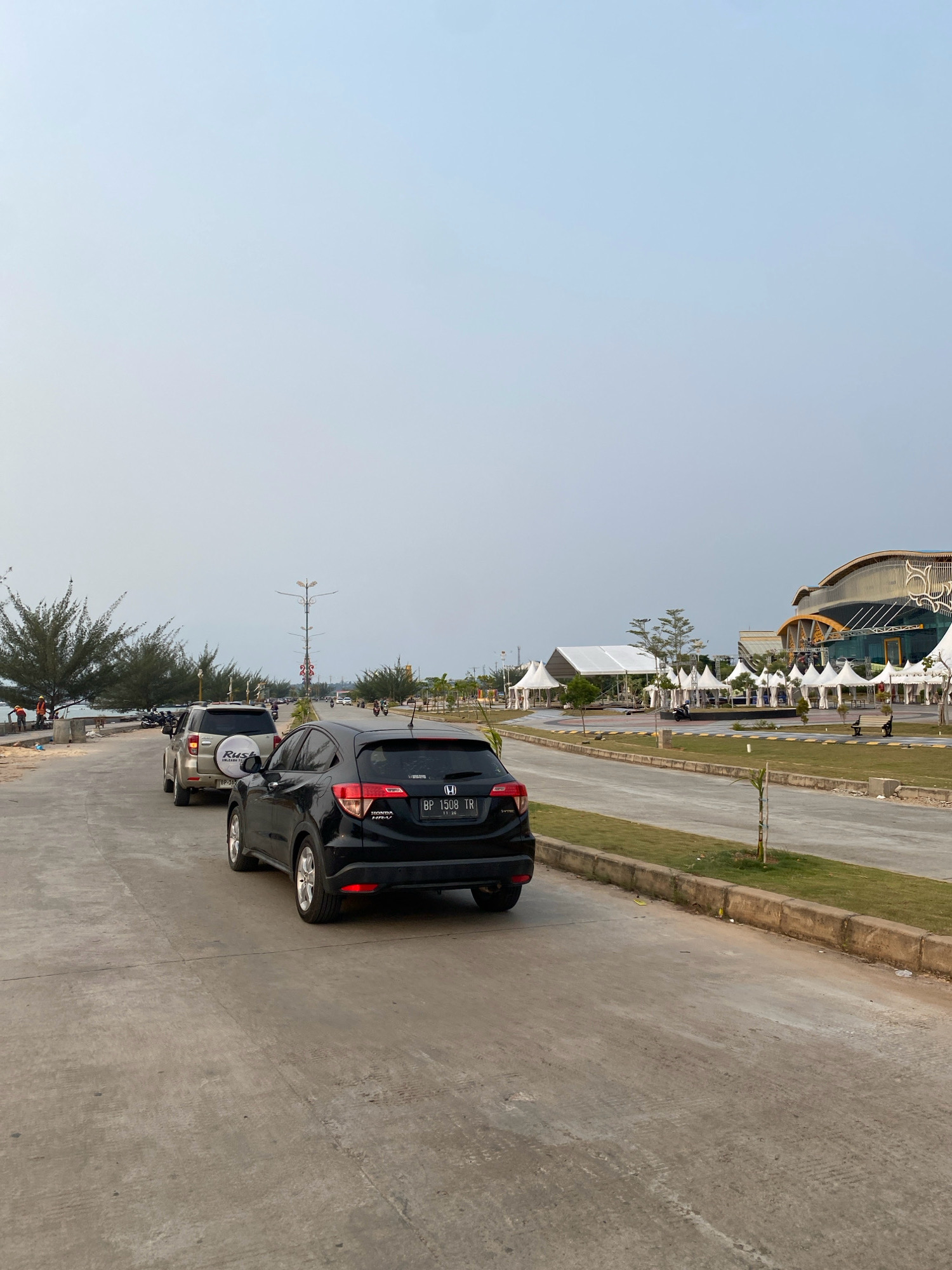 This image depicts a street scene in the city of Tanjung Pinang, Indonesia, likely near a waterfront or event venue. The foreground features a black Honda HR-V with license plate BP 1508 TR, positioned on a paved road. Behind it, a silver SUV with a spare tire mounted on the back is also visible, along with other vehicles further down the road. The road is bordered by a grass verge and a concrete curb. In the background, there are several white tents set up, suggesting an outdoor event or market. A large, modern building with a distinctive architectural design is situated to the right, possibly a convention center or entertainment venue. Trees, including pine-like varieties, line the road on the left side, with a glimpse of water and a few individuals working on what appears to be construction or landscaping near the water's edge. The sky is overcast and light blue, indicating daytime conditions.