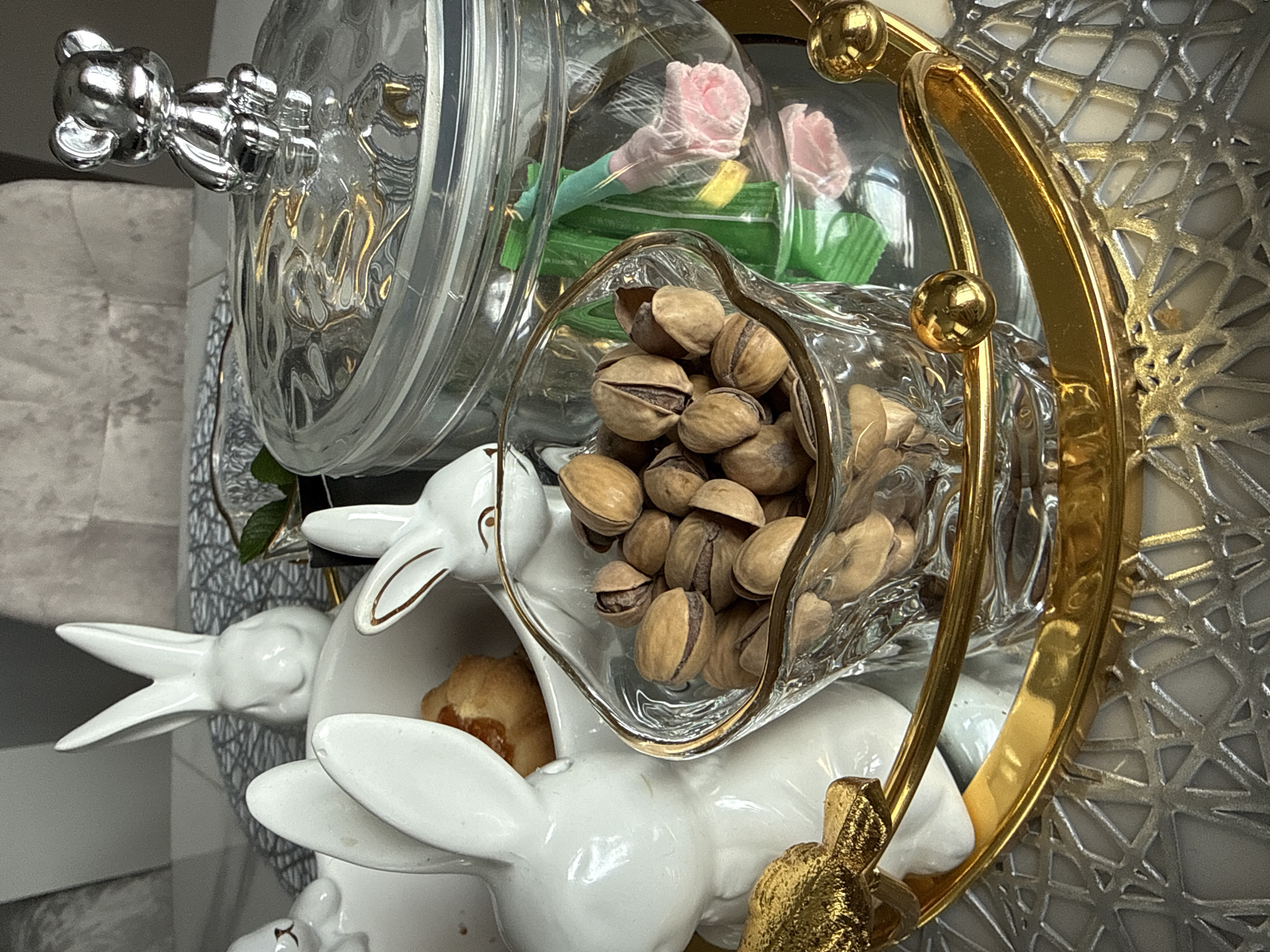 The image shows a decorative centerpiece. It consists of a stack of white ceramic rabbits supporting a tiered glass container with gold trim. The top tier contains artificial pink flowers. The second tier contains pistachios. The bottom tier has a pastry. The scene is set against a silver decorative backdrop. The location is not discernible from the image.