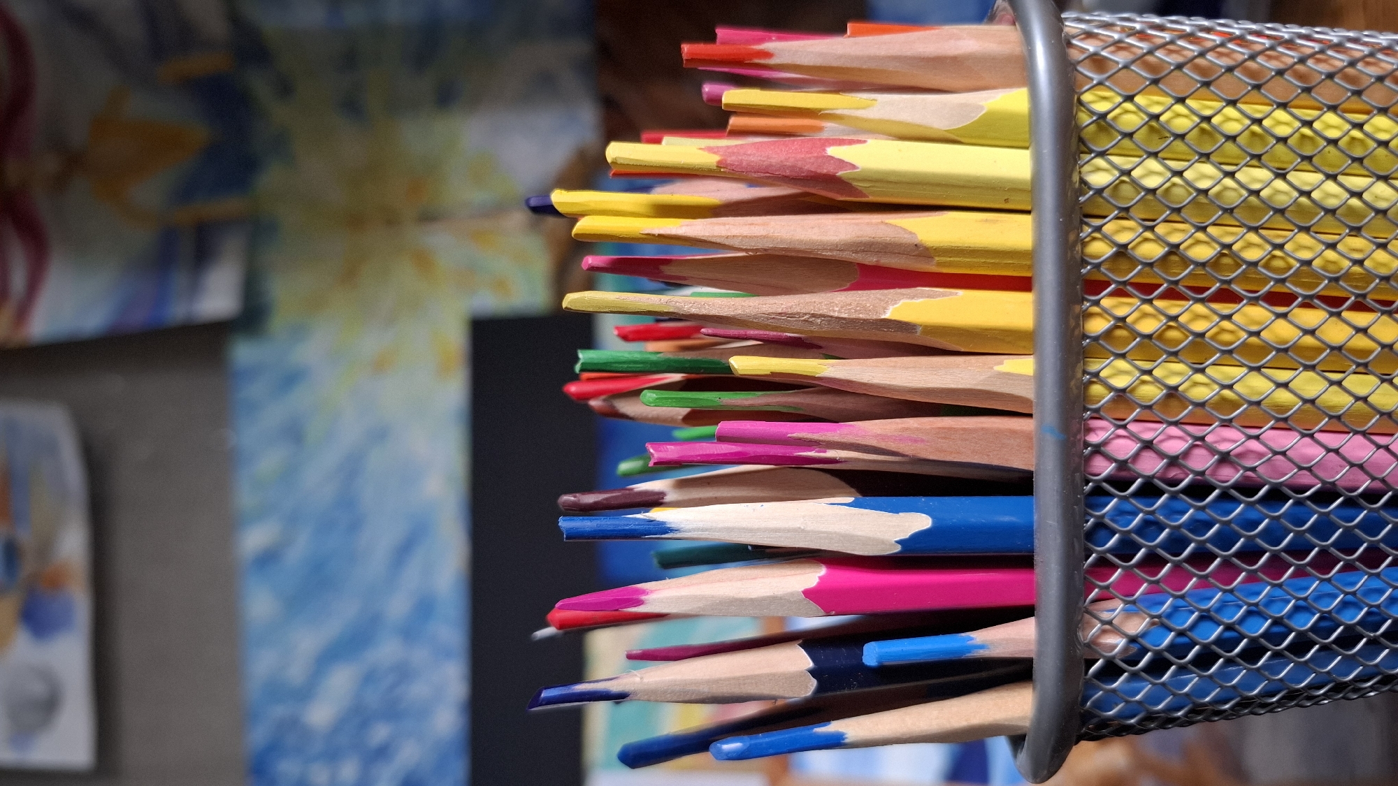 A close-up, eye-level photograph captured in Novaya Vasil'evka, Russia, depicts a metal mesh pencil holder densely packed with numerous sharpened colored pencils. The holder is silver-gray, featuring a regular diamond-patterned mesh grid, with a smooth, slightly thicker rim at the top. The pencils inside are upright, showcasing a vibrant array of colors, predominantly blue, magenta/pink, yellow, red, and green. Many pencils are sharply pointed, revealing the colored lead tips and the light-toned natural wood underneath the paint. The background is out of focus, indistinctly showing what appears to be various artworks or colored surfaces, including areas of blue and yellow, indicating an indoor, likely artistic or creative, environment. Natural light illuminates the scene, creating soft highlights on the pencil tips and the mesh. No individuals are present in the image.