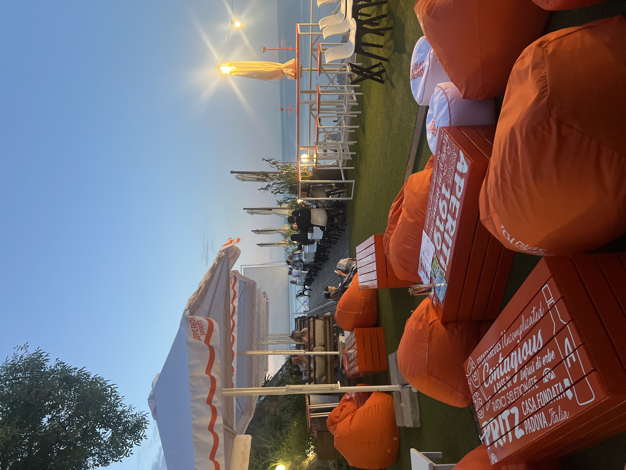 The image depicts an outdoor seating area at a waterfront establishment in Odesa, Ukraine, likely during twilight. The scene features an arrangement of orange beanbag chairs and low, bright orange wooden tables with white lettering, possibly a brand name, scattered across a green, artificial turf surface. White umbrellas with orange trim provide shade for some areas. In the background, a long table with white chairs is set up, and several patrons are seated, facing a body of water that appears to be the Black Sea. A row of lights and a tall, illuminated umbrella-like structure are visible, indicating a lively evening atmosphere. The sky is a clear, soft blue transitioning to a lighter hue near the horizon, with the sun having already set. The overall impression is a relaxed, open-air lounge or restaurant by the sea.