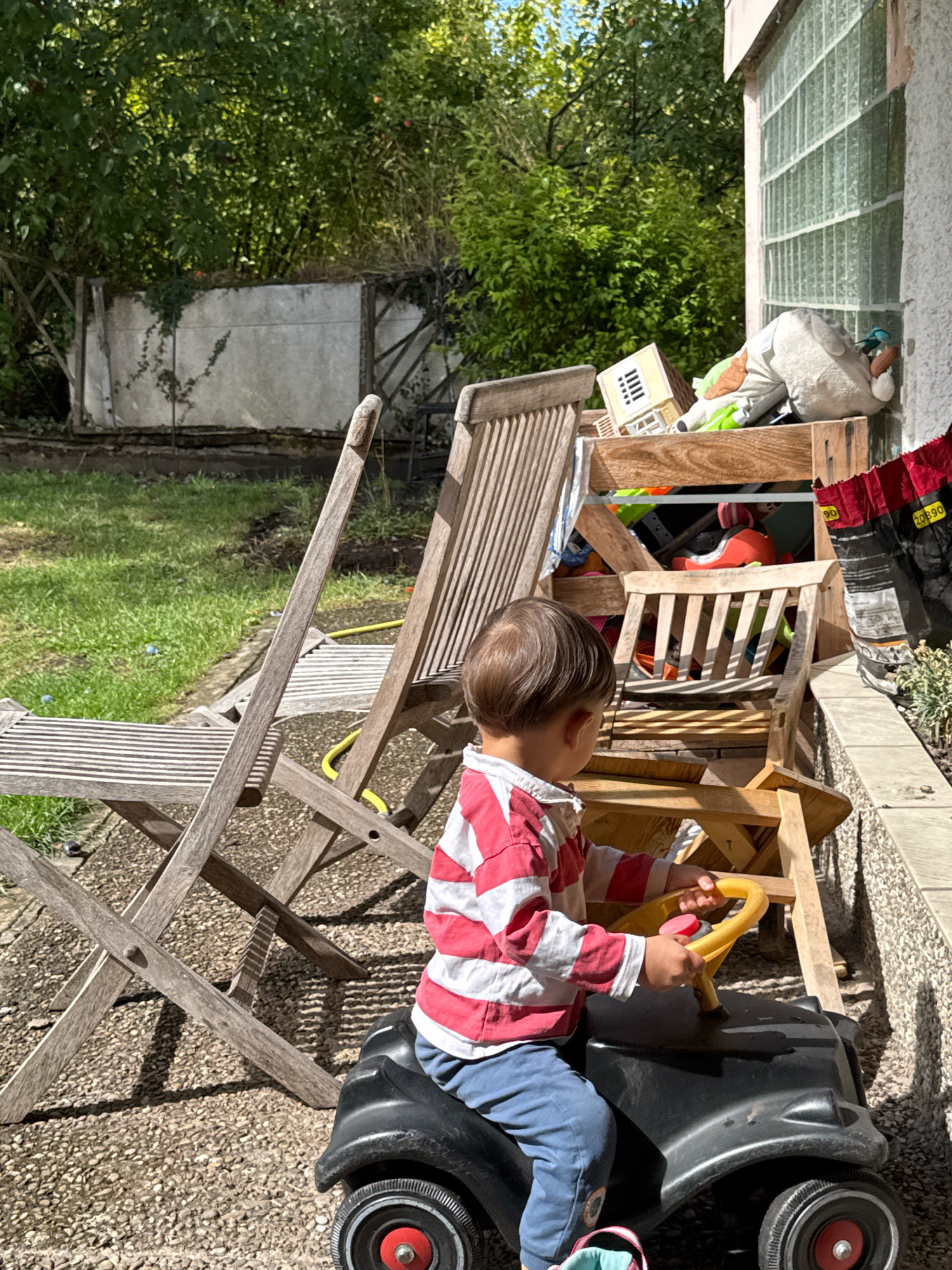 A young child wearing a red and white striped shirt and blue pants sits in a black toy car with a yellow steering wheel. The child is positioned on a gravel surface in a backyard. In the background, there are several wooden chairs, a stack of various items in a wooden crate, and lush green trees. A portion of a building with glass block windows is visible on the right side of the frame. The scene is illuminated by natural sunlight, casting shadows from the chairs and other objects. The location is identified as a backyard in Überherrn, Germany.