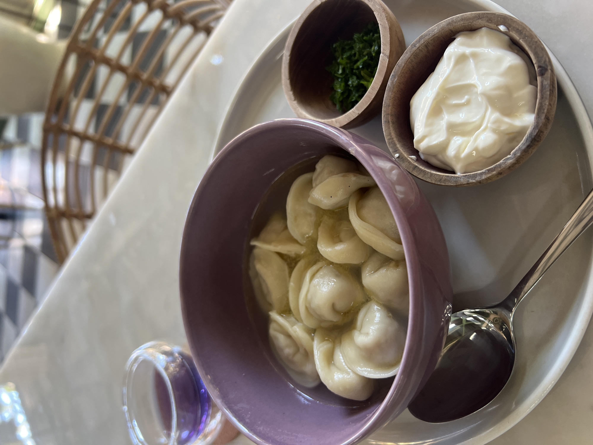 The image depicts a serving of pelmeni, a type of dumpling, in a purple bowl filled with broth. The bowl rests on a white plate. To the side of the bowl are two small wooden bowls, one containing chopped green herbs, likely parsley or dill, and the other filled with a white, creamy substance, possibly sour cream or yogurt. A silver spoon also lies on the plate. A small glass container filled with a purple liquid is seen near the bowl of pelmeni. The background shows a woven wicker object and a glimpse of black and white checkered flooring. The location is Sukawati, Indonesia.