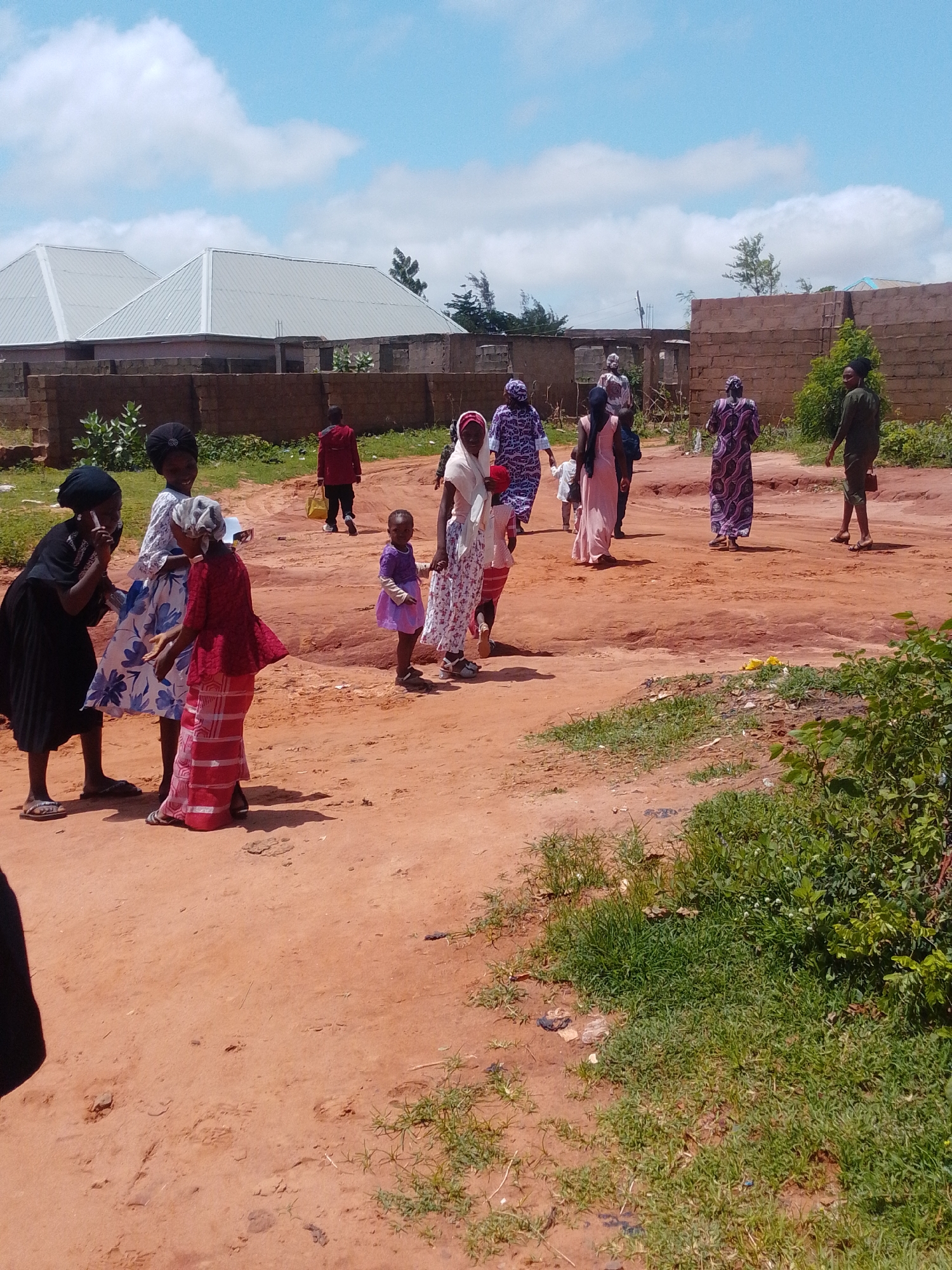 In Tumfure, Nigeria, a group of people, primarily women and children, are gathered in an outdoor, dusty area. Two buildings with light-colored, sloped roofs are visible in the background, behind a low, brick wall. Some of the women are wearing traditional attire, including headscarves and long dresses. One woman in the foreground, wearing a black dress, appears to be looking at her phone, while another woman in a white and blue patterned dress stands next to her. A young child in a purple dress is walking in the middle of the scene. Further back, several other individuals, including a man in a green shirt and a woman in a dark patterned dress, are walking. The ground is covered in reddish-brown dirt, with patches of dry grass and some scattered debris. The sky is partly cloudy with a bright blue hue.