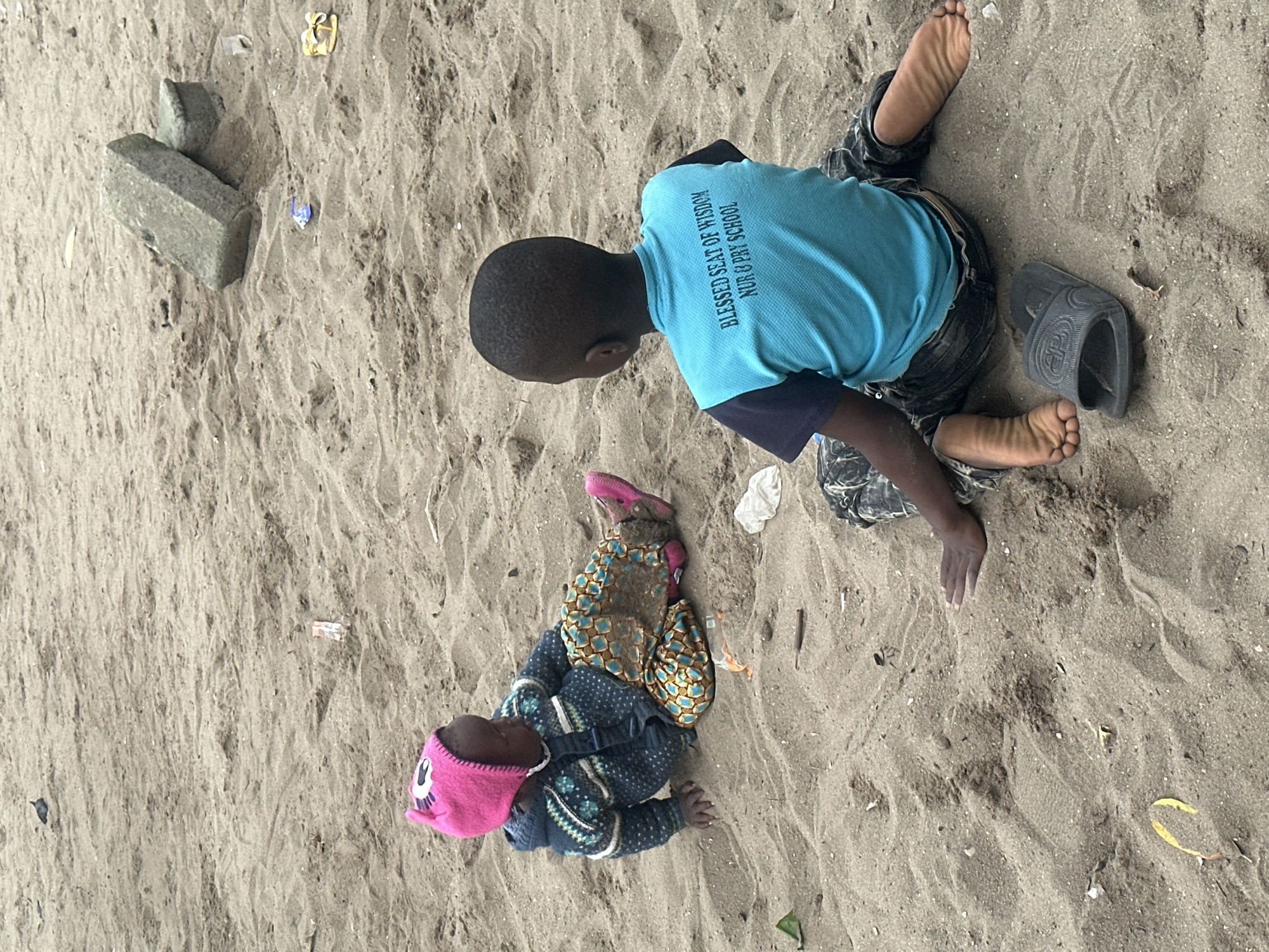 The image depicts two Black children sitting on a sandy ground in what appears to be an outdoor area. The child on the left, facing to the right, is wearing a pink hooded hat with an owl design, a patterned sweater, and patterned pants with pink shoes. The child on the right, facing away from the camera, is wearing a blue t-shirt with text that reads "BLESSED SEAT OF WISDOM NUR & PRY SCHOOL" and dark patterned pants. The child on the right has a black sandal next to their left foot. The sandy environment shows signs of debris, including what looks like plastic wrappers and other small litter. In the background, two large rectangular stones are visible, one stacked on top of the other. The setting is likely a public area or a beach, possibly in a less developed part of Badagry, Nigeria, given the educational institution mentioned on the t-shirt.