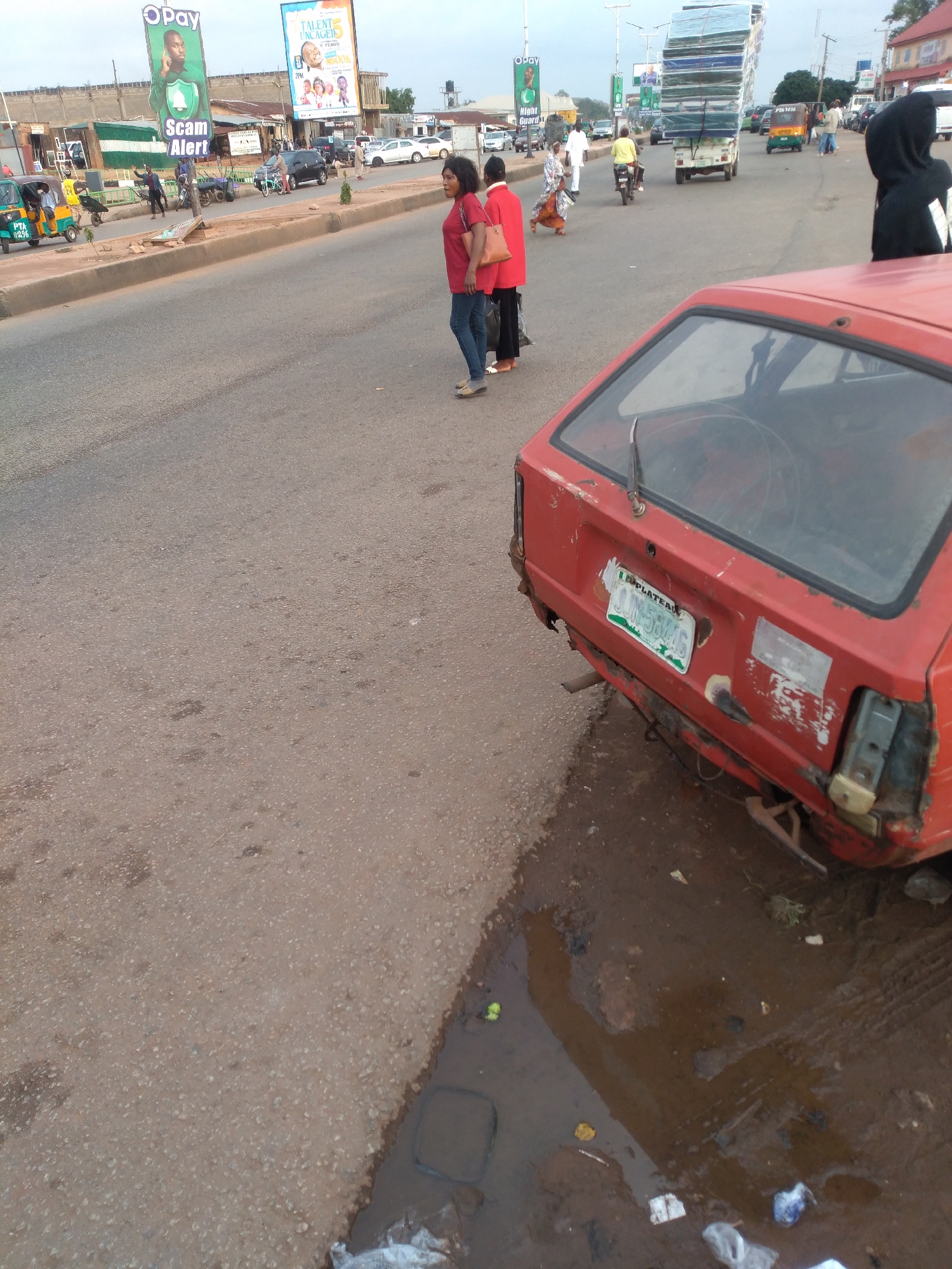 The image depicts a street scene in Gura Top, Nigeria, with a red car in the foreground, showing signs of damage and disrepair. The car's license plate, partially visible, reads "JUIN 58146" and is affixed to the rear. The surrounding area is a paved road with debris, including what appears to be trash and a puddle of water. In the background, several people are visible, including a woman in a red top and jeans walking towards the left, and another person in a black hoodie standing on the right. Other individuals are seen near a tuk-tuk and on motorcycles, suggesting everyday street activity. Buildings and billboards are present in the distance, including an "OPay" advertisement. The sky is overcast, indicating diffused lighting conditions.
