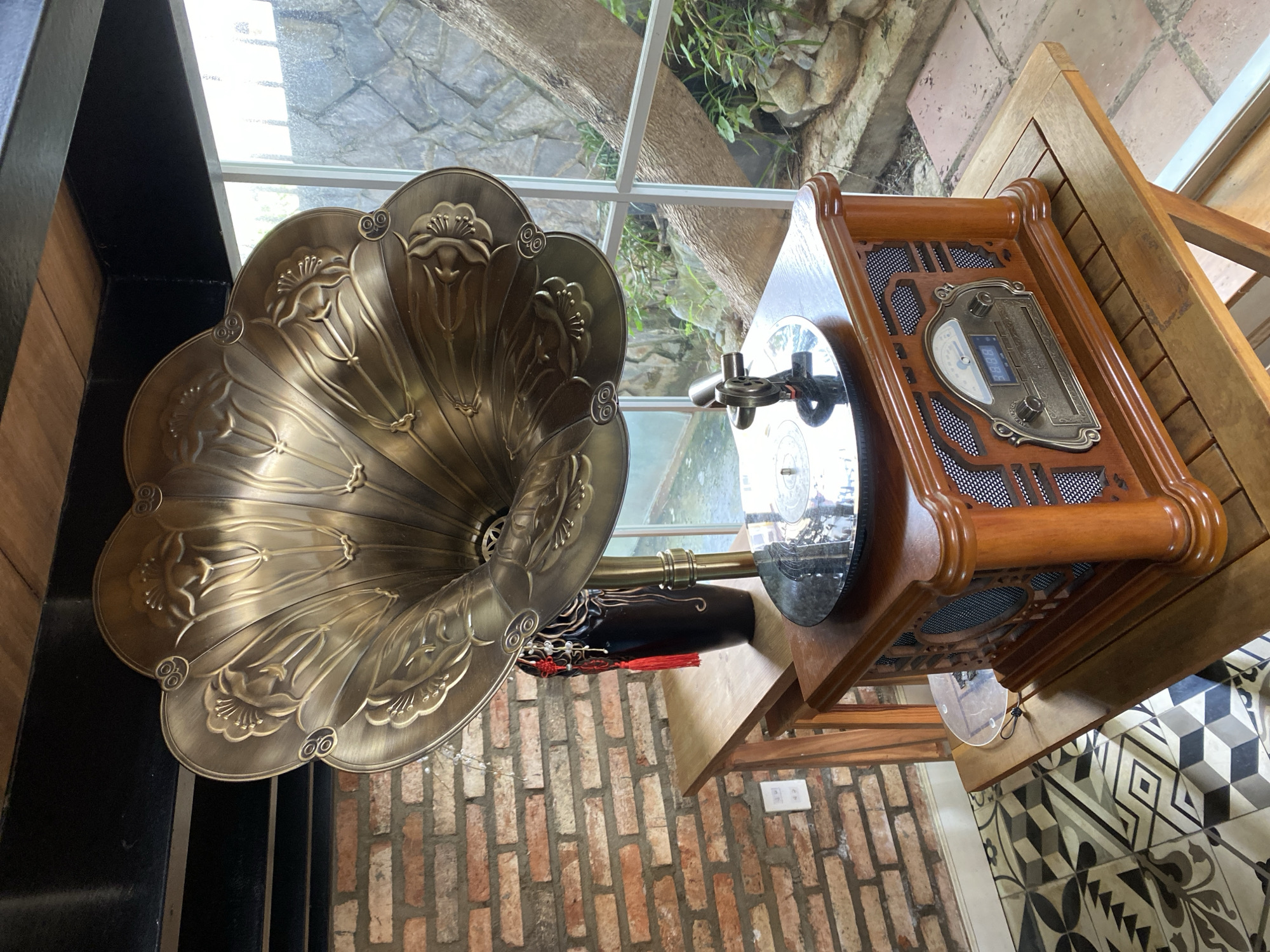 The image shows an antique-style record player on a wooden table. The record player is made of wood and has a brass horn. It is placed in a room with a brick wall and a window that looks out onto a garden. The flooring is tiled with a geometric pattern. The record player appears to be functioning, with a record on the turntable and the needle down. The overall scene suggests a cozy and nostalgic atmosphere. Based on the visual cues of the brickwork, the style of the furniture, and the presence of lush greenery visible through the window, this setting is consistent with a cafe or a homestay in Da Lat, Vietnam, known for its colonial architecture and natural beauty.
