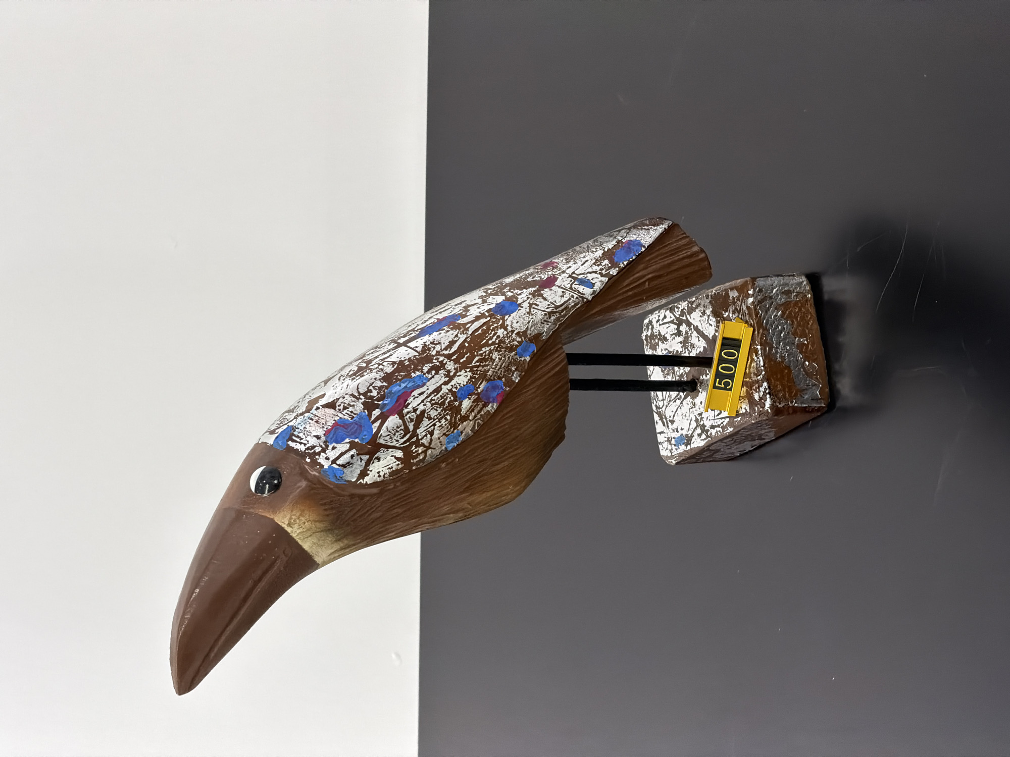 The image displays a decorative bird figurine. The figurine is primarily brown with a long, pointed beak and a single black eye. The body of the bird is adorned with white, blue, and pink paint splatters, giving it a textured and artistic appearance. The bird is mounted on two thin black rods, which are attached to a rectangular base. This base is also decorated with a brown and white textured pattern. A small yellow tag with the number "500" printed in black is affixed to the front of the base. The figurine is positioned on a dark, reflective surface, possibly a table. The background consists of a white wall above a dark gray wall. There are no people or animals present in the image. The setting appears to be an indoor display or retail environment, but no specific location in Minsk, Belarus, can be identified from the visual information provided.