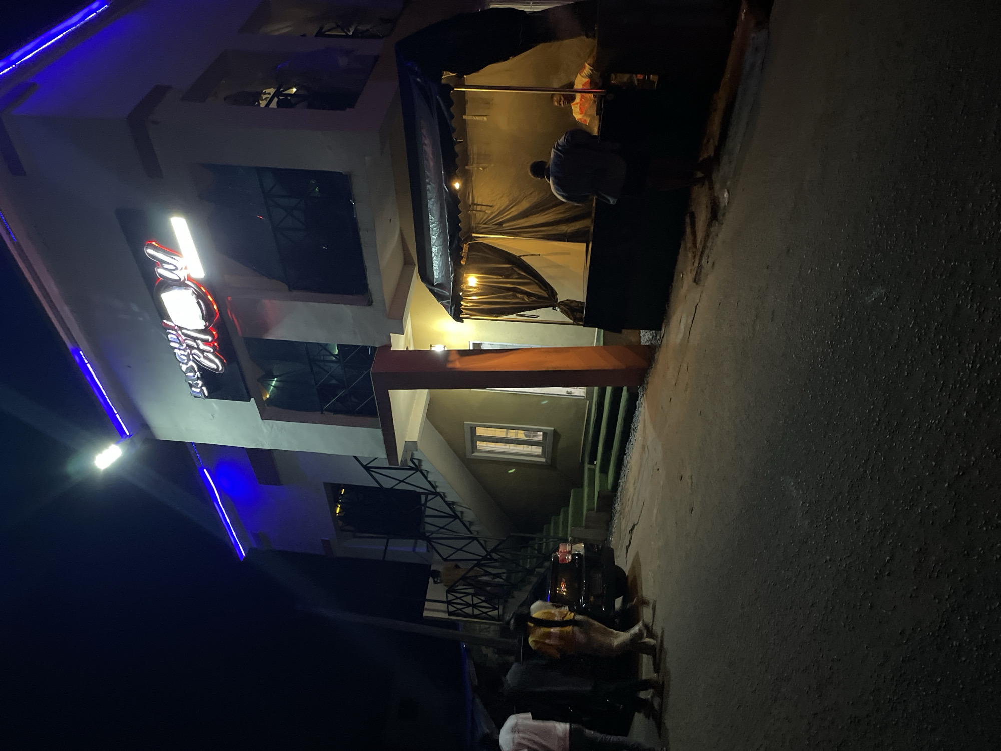 This nighttime image captures a street scene in Jos, Nigeria, featuring a two-story commercial building, a vendor stall, and six discernible individuals.
The two-story building on the left has light-colored walls. Its upper level is outlined by blue LED strip lighting along the eaves, and a bright white floodlight is mounted on the upper left corner. A prominent illuminated sign on the second floor reads "SAUDI CHOW," with "CHOW" displayed in red neon-style lettering, accompanied by a bright white rectangular light. A staircase with dark railings, featuring steps covered in green artificial turf, leads to the second floor from the ground level on the left side. A dark-colored SUV is parked at the base of these stairs.
To the right, a dark vendor stall or kiosk stands, covered by a black tarp roof and dark fabric sides. It is illuminated internally by warm-toned hanging lights.
A total of six individuals are present. Four people are observed in the vicinity of the parked SUV and the building's staircase: one partially visible individual in a white shirt on the far left; another in a dark shirt near the SUV; a person in a yellow and orange patterned shirt walking away from the car towards the right; and an individual in a light-colored shirt ascending the green-covered stairs. At the vendor stall, one person in a dark blue or grey shirt stands with their back to the camera, facing the stall, interacting with a seated individual inside the stall, who wears a light-colored, patterned shirt.
The ground surface consists of dark, textured asphalt pavement, showing visible cracks in the foreground. The scene is primarily lit by the artificial light sources from the building and the vendor stall.