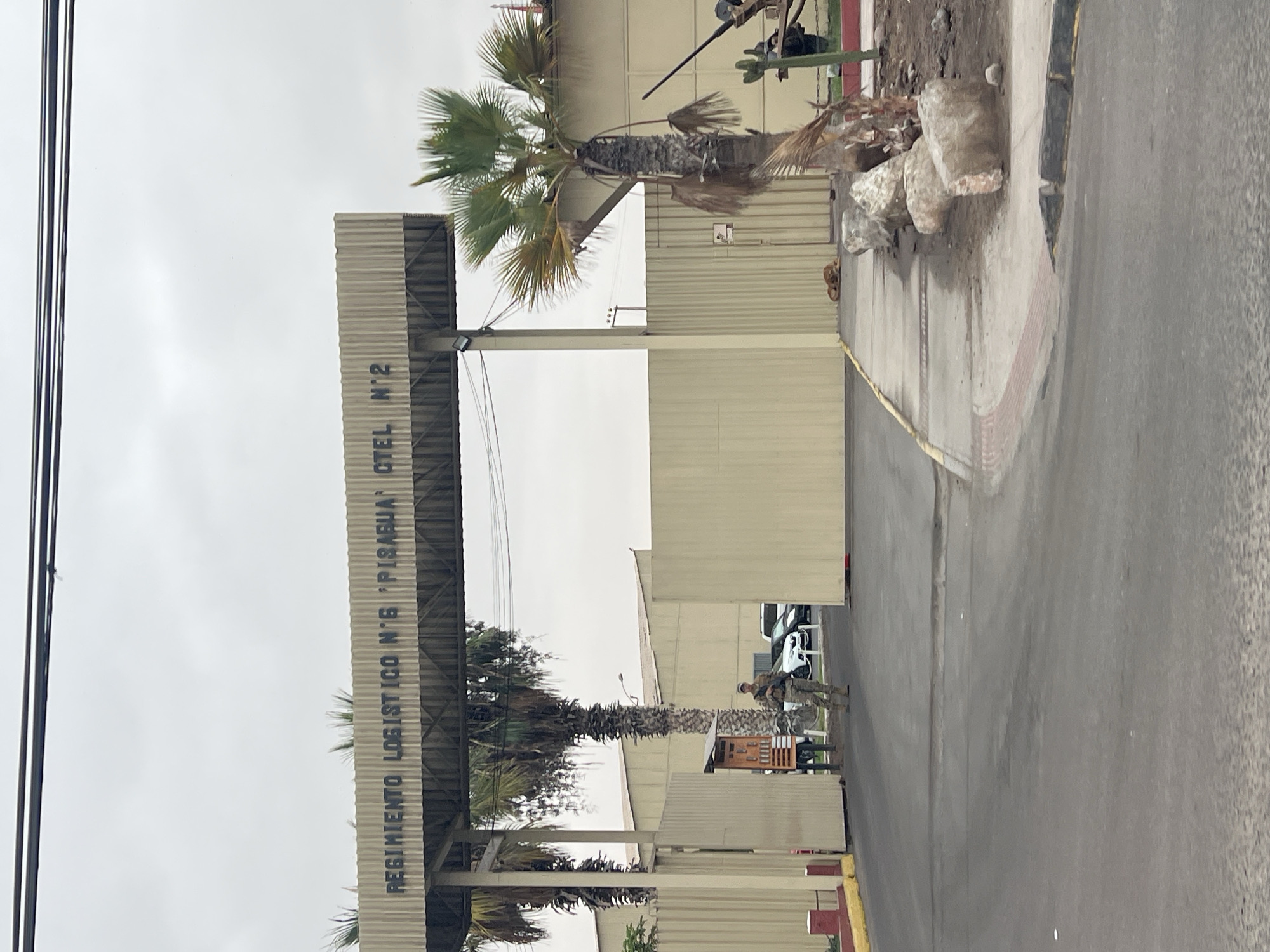 The image depicts the entrance to the "Regimiento Logistico N.6 Pisagua Hotel N.2" in Arica, Chile. A soldier in military fatigues, carrying a rifle, stands guard at the entrance. A guard booth is near the soldier. Vehicles are visible inside the compound. The entrance has a metal awning with the regiment's name printed in dark letters. Palm trees flank the entrance. The weather appears overcast. The road is asphalt. A concrete sidewalk with a yellow tactile paving strip is seen on the right.