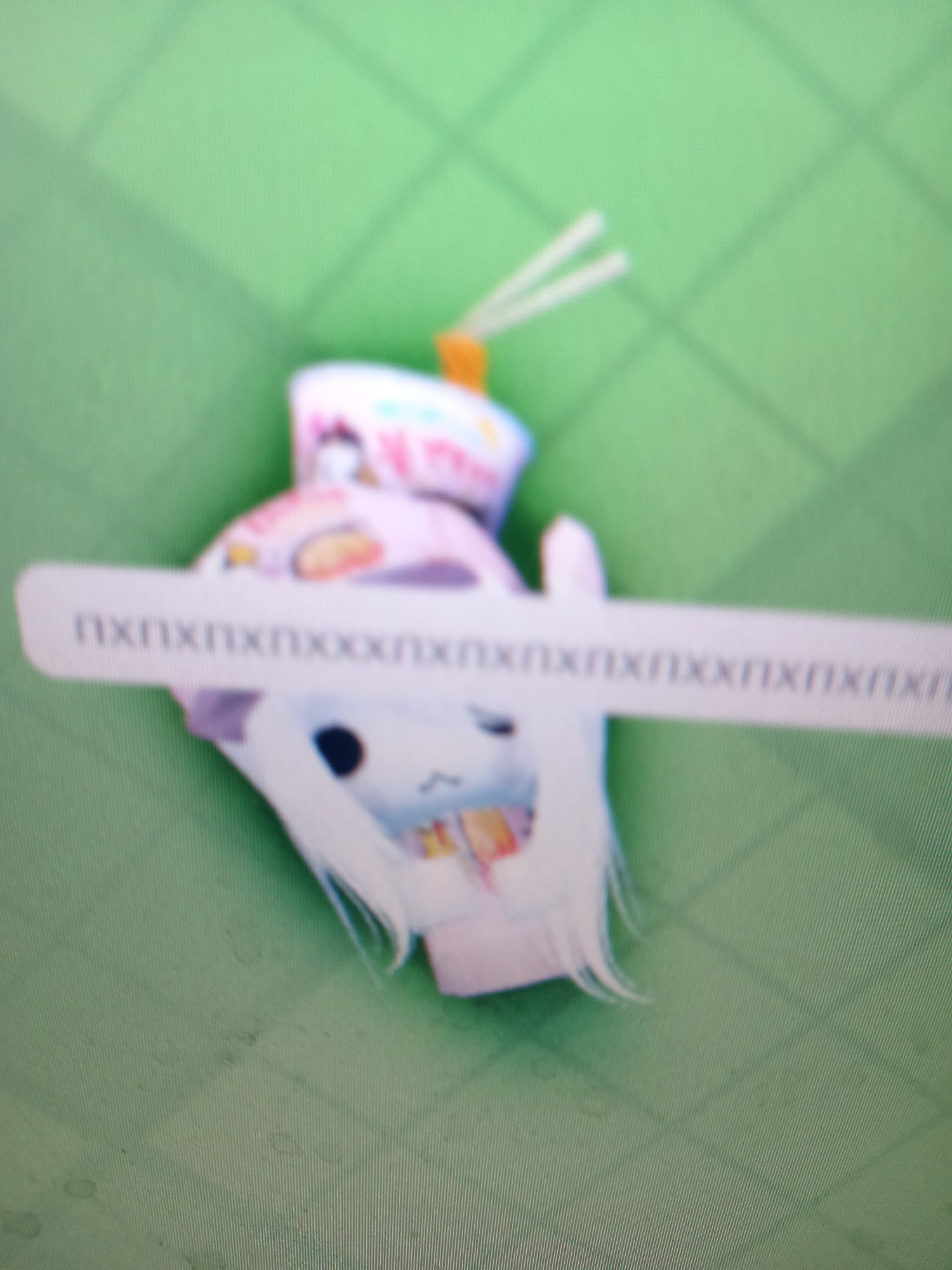 The image shows a blurry, overhead shot of a stuffed toy, possibly a cat or rabbit, with white hair-like strands and a pink and white hat, on a green surface with a grid pattern. A white sticker or label with the text "nxnxnxnxnxxnxnxnxnxnxnxnxnxnx" is partially visible, overlaid on the toy. The location in Rudny, Kazakhstan, is not discernible from the provided image.