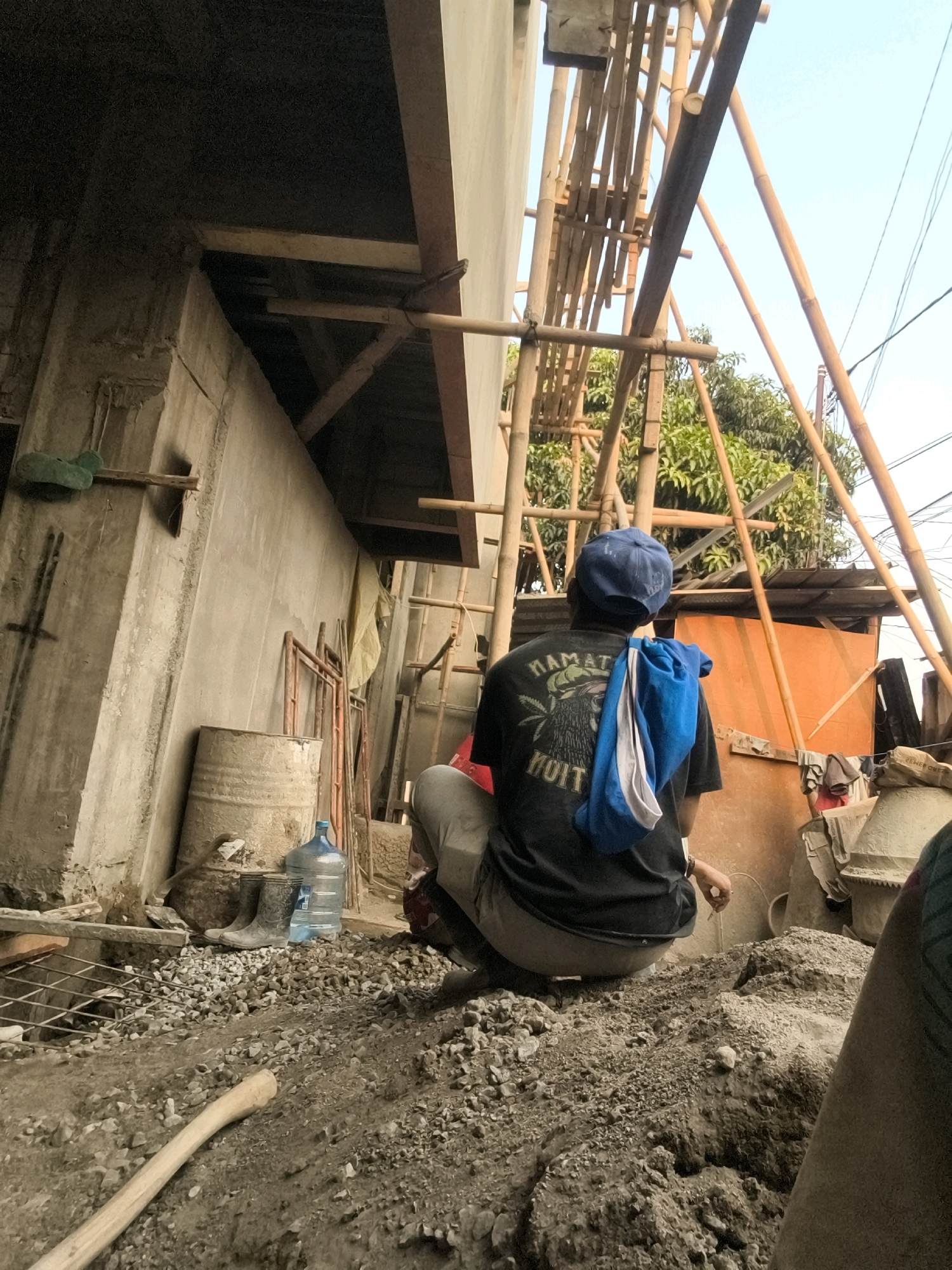 The image depicts an outdoor construction site in Bekasi, Indonesia. A man wearing a blue baseball cap and a black t-shirt with graphics on the back is squatting on a pile of construction debris. A blue and white cloth is draped over his shoulders. In the background, bamboo scaffolding is erected against a building under construction. Various construction materials and equipment are visible, including a metal barrel, a large water jug, a pair of rubber boots, a shovel handle, and what appears to be a concrete mixer. The environment suggests a typical urban construction scene in the city.