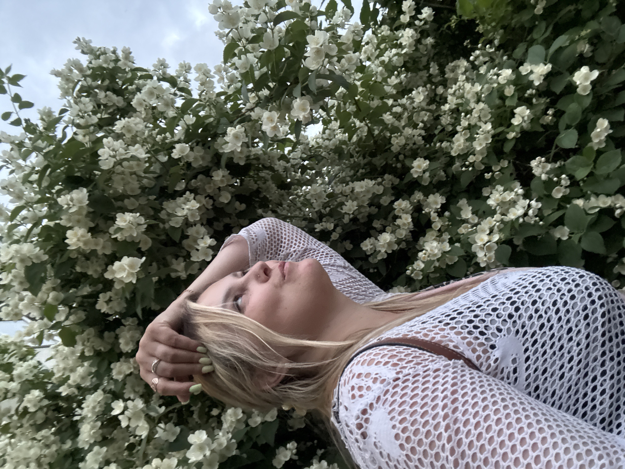 A young woman with blonde hair and light skin, wearing a white mesh top and a black undergarment, is captured in profile against a dense backdrop of white jasmine flowers and green leaves. Her right arm is raised, with her hand resting on her head, adorned with two rings. Her left arm is partially visible, also covered by the mesh top. She is looking upwards and to the right with a thoughtful expression. The background is dominated by the white blossoms, with a cloudy sky visible in the upper right corner. The scene suggests an outdoor setting, possibly a park or garden, given the abundant flowering shrubbery, and could be located in Chaikovsky, Russia, a city known for its natural beauty and parks.