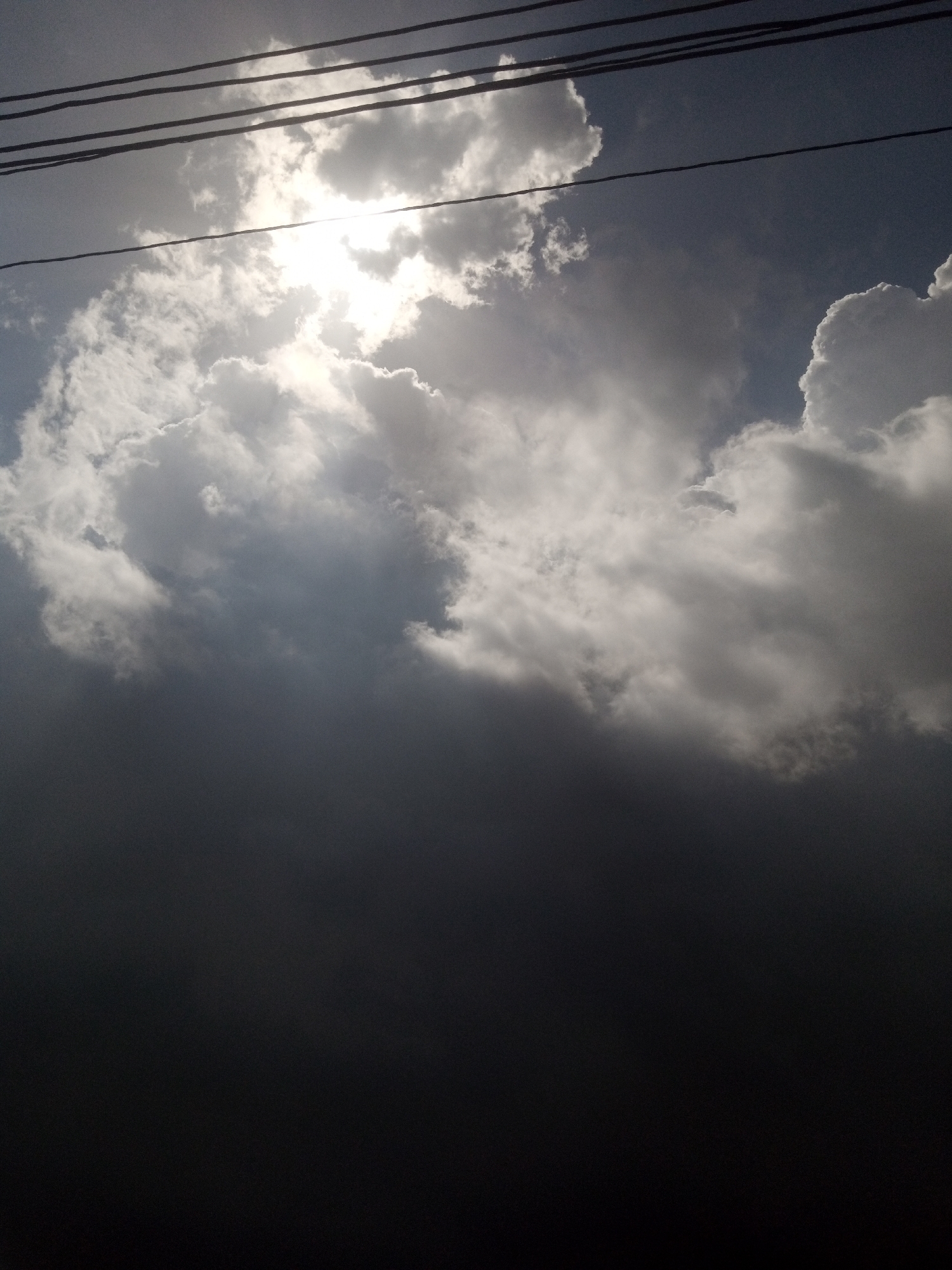 The image shows a dark sky with scattered clouds. The sun is partially obscured by clouds, creating a bright glow in the upper center of the frame. Several power lines stretch horizontally across the top of the image. The bottom half of the image is very dark and featureless, suggesting it might be an overexposed area or a dark foreground element that is not clearly defined. The overall impression is of a dramatic and somewhat ominous sky.