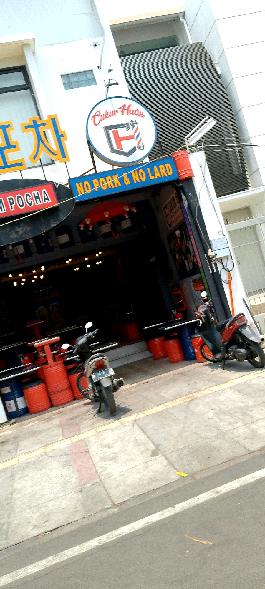 This is a street-level, slightly tilted, full-shot image of a barber shop named "CUKUR HADE" located in Bandung City, Indonesia. The shop has a distinct blue sign with red and white lettering that reads "NO PORK & NO LARD" above the entrance. To the left of the barber shop sign, there are large, yellow, stylized Korean characters on a blue background. In front of the shop, there are several large red plastic barrels and two blue barrels, along with some tables and chairs that are not fully visible. Two motorcycles are parked on the sidewalk in front of the shop, one silver and one red. The sidewalk is paved with concrete tiles and has a yellow tactile paving strip along its edge. The street in front of the sidewalk appears to be asphalt with a white line. The building behind the shop is modern, with white and grey paneling and a large window. There are overhead wires and a lamppost visible near the building. The sky is clear and bright, suggesting it is daytime. No people are clearly visible in the image.