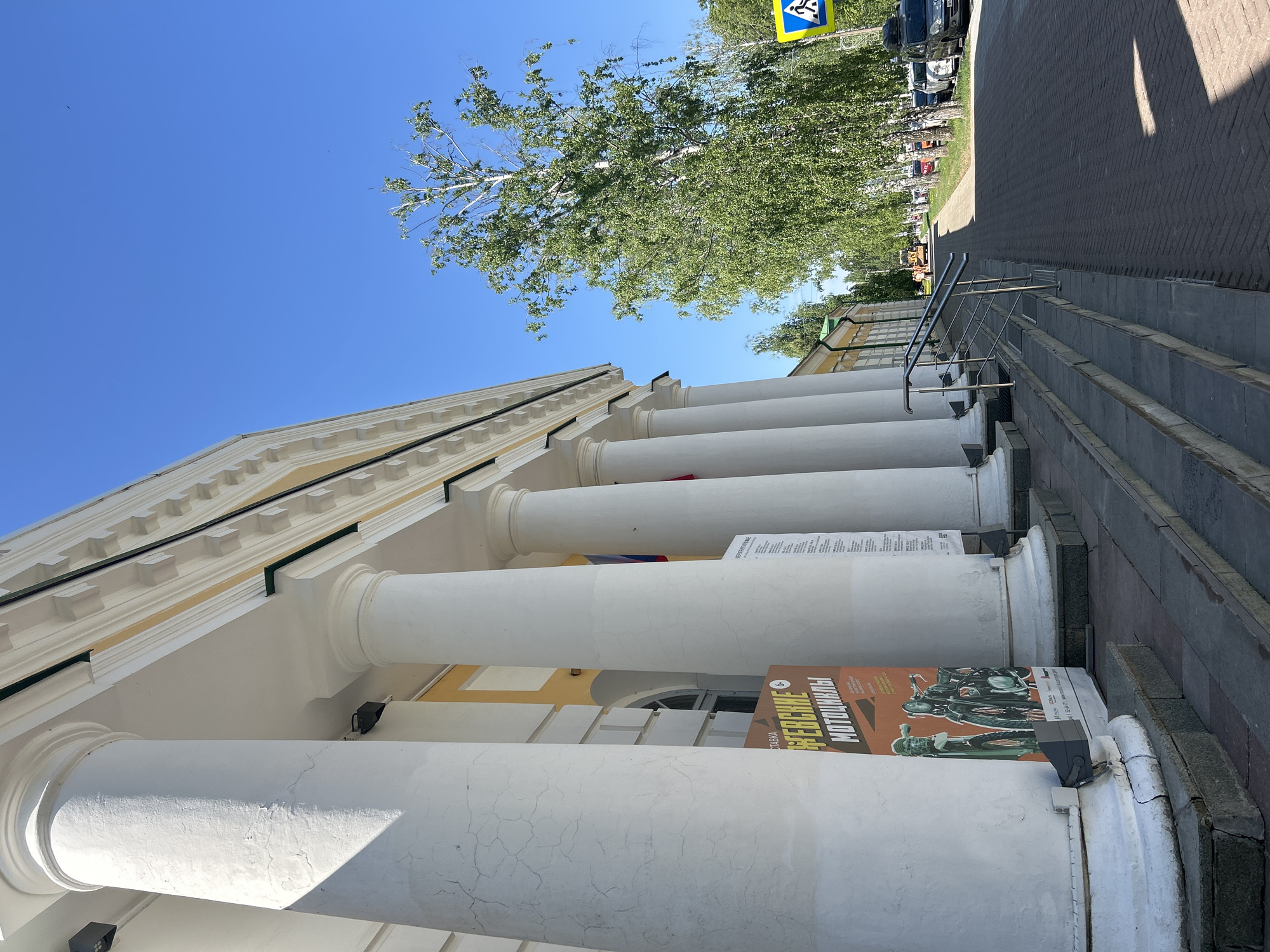 The image shows a building with white pillars in Izhevsk, Russia. The building has a yellow facade and a row of white pillars. There is a poster advertising an exhibition of vintage motorcycles leaning against one of the pillars. A staircase with metal handrails leads up to the building. There is a sidewalk and some parked cars in the background. A tree with green leaves is visible above the building. The sky is clear and blue.