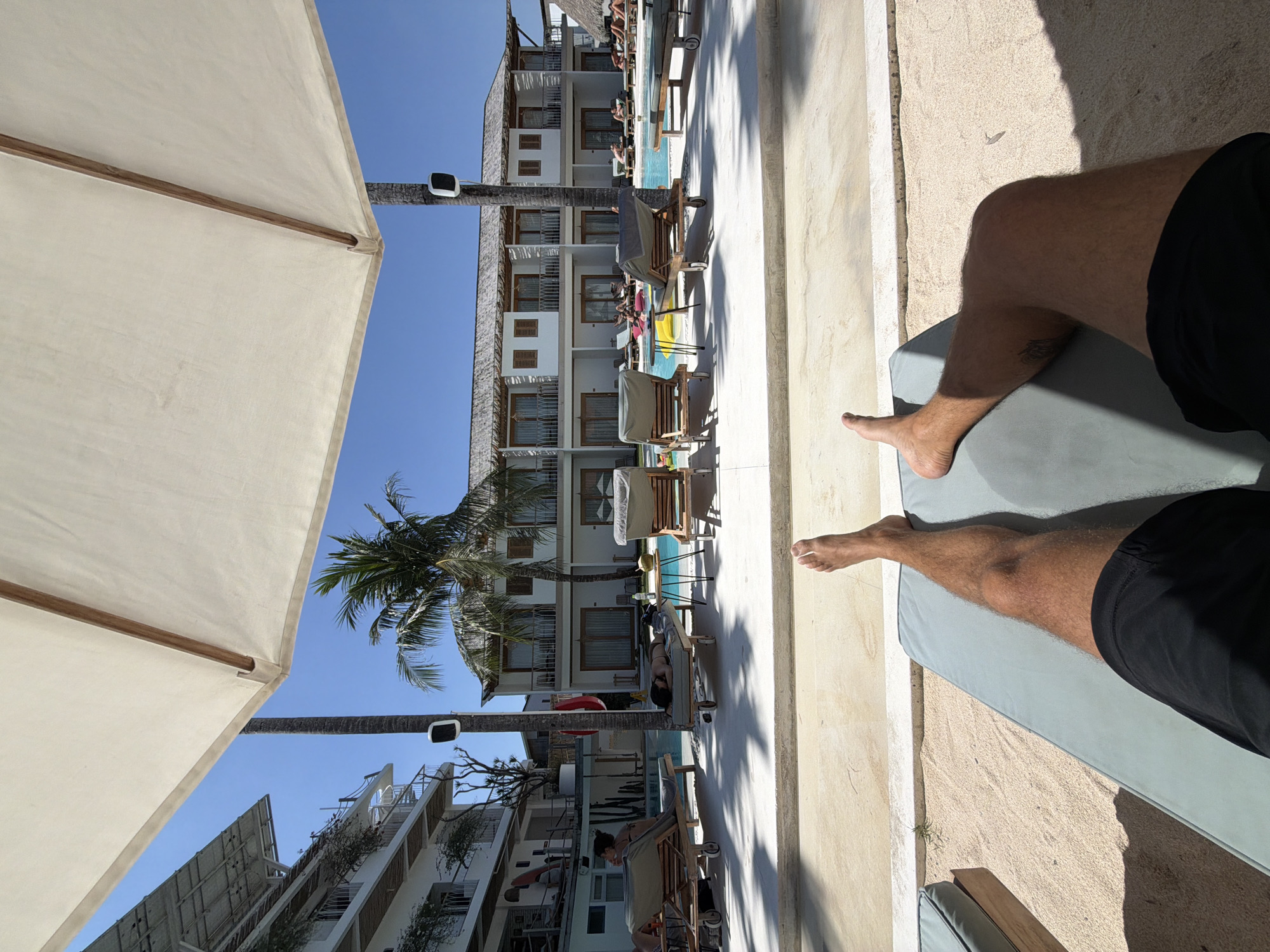 A first-person perspective shot captures a person relaxing on a lounge chair under a large white umbrella. The scene is set at a resort in North Kuta, Indonesia. In the background, there is a swimming pool, several lounge chairs, palm trees, and a multi-story hotel building with balconies. The sky is clear and blue.