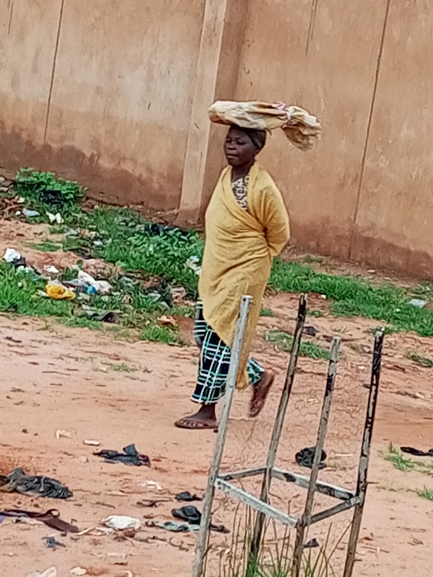 In Tumfure, Nigeria, a woman walks along a dirt path beside a wall. She wears a yellow headscarf and a yellow wrapper over a patterned skirt. On her head, she balances a large bundle wrapped in paper. The ground is covered in scattered debris, including plastic bags, fabric scraps, and other unidentifiable refuse. Green vegetation grows in patches throughout the area. In the foreground, a rusted metal structure, possibly part of a discarded chair, stands on the path. The wall behind the woman is rough and textured, with signs of moisture damage near the ground. The overall scene depicts a gritty urban environment with visible signs of neglect and poverty.