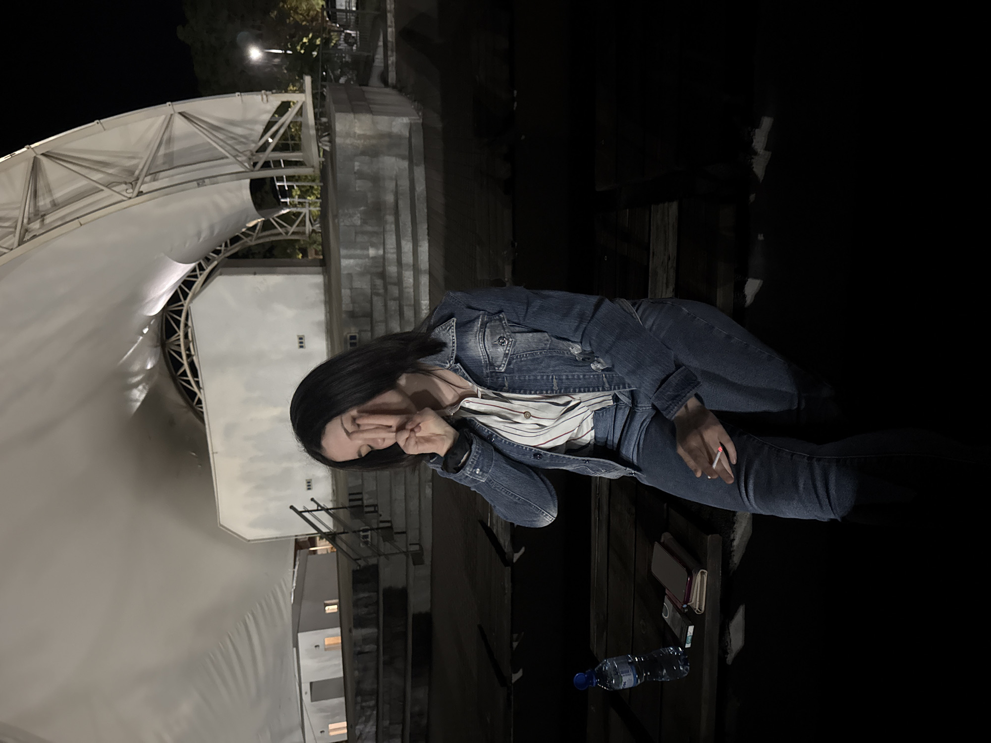 The image shows a woman seated on a wooden surface, likely a bench, at night. She is wearing a denim jacket and jeans, and a striped shirt. She holds a lit cigarette in her right hand and is covering her eyes with her left hand. Next to her is a bottle of water, a phone, and what appears to be a notebook. The background features a large white structure, potentially an amphitheater or outdoor stage, indicating an urban setting. The location is in the city of Unknown, Unknown.