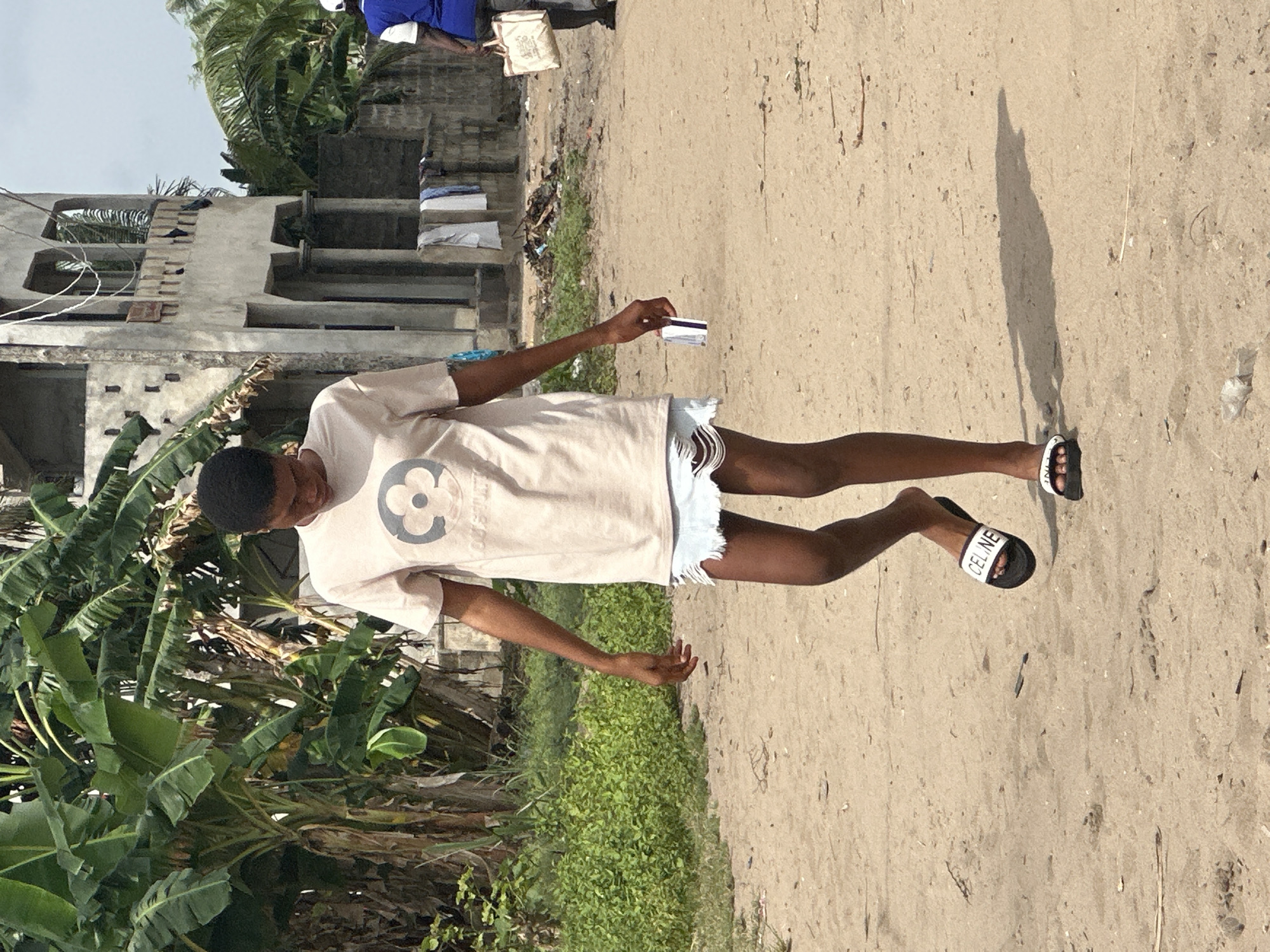 A young person with short dark hair and dark skin stands on a sandy or dirt path under bright daylight. They are wearing a light-colored t-shirt adorned with a dark, stylized flower-like logo, and light-colored ripped denim shorts. On their feet are black slide sandals, one of which clearly displays "CELINE" in white text on its strap. In their right hand, they hold a small, white plastic card with a black magnetic stripe, resembling a credit or identification card. Their shadow is cast long and distinct on the ground, indicating strong sunlight.
The setting is outdoors in what appears to be a tropical or semi-tropical environment. To the left, lush, dense green vegetation, including large-leaf plants possibly banana trees, lines the path. In the background, partially constructed or unfinished concrete buildings with exposed rebar and brickwork are visible, suggesting a developing area. The sky above is clear. The scene captures a moment of an individual posing or standing, holding a card, amidst a natural and partially constructed landscape, typical of the provided location context of Badagry, Nigeria.