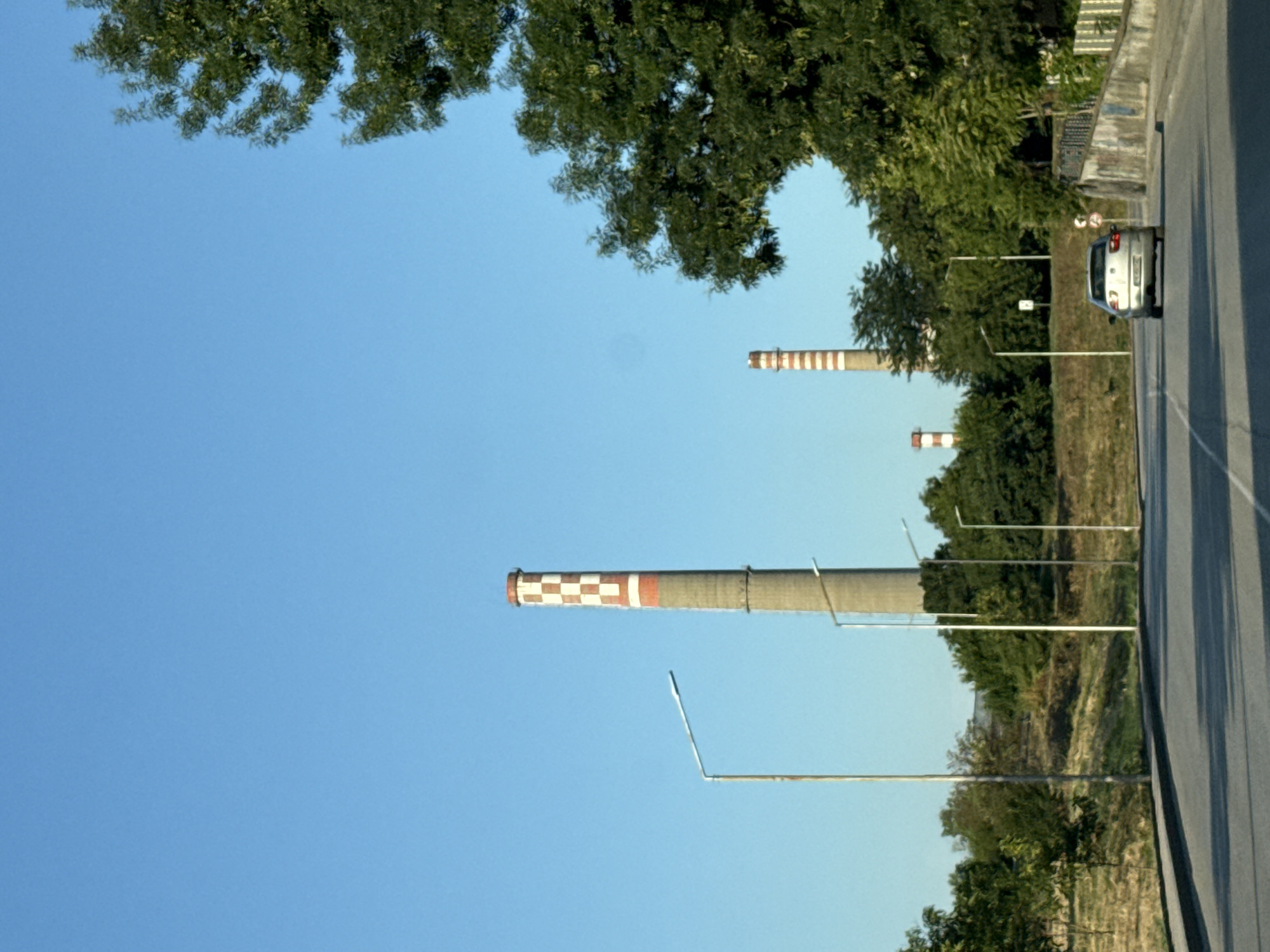 The image captures a view of smokestacks in the city of Pleven, Bulgaria, likely part of an industrial facility, possibly a power plant. In the foreground, a two-lane asphalt road is visible, with a light-colored car driving away from the viewer. The car is centered in the right lane. The road is bordered on the left by a grassy, uneven embankment with sparse vegetation and several tall, slender streetlights. On the right side of the road, a textured concrete wall with graffiti runs along the edge, leading to a fence. A large, leafy green tree dominates the right side of the frame, partially obscuring the sky. In the background, two prominent smokestacks stand tall against a clear blue sky. The closer smokestack is taller, with distinct red and white checkered bands near its top, and a gray concrete base. Further in the distance, a second, shorter smokestack with similar red and white banding is visible, surrounded by more trees and foliage. The overall scene suggests a semi-urban industrial landscape on a clear, bright day.