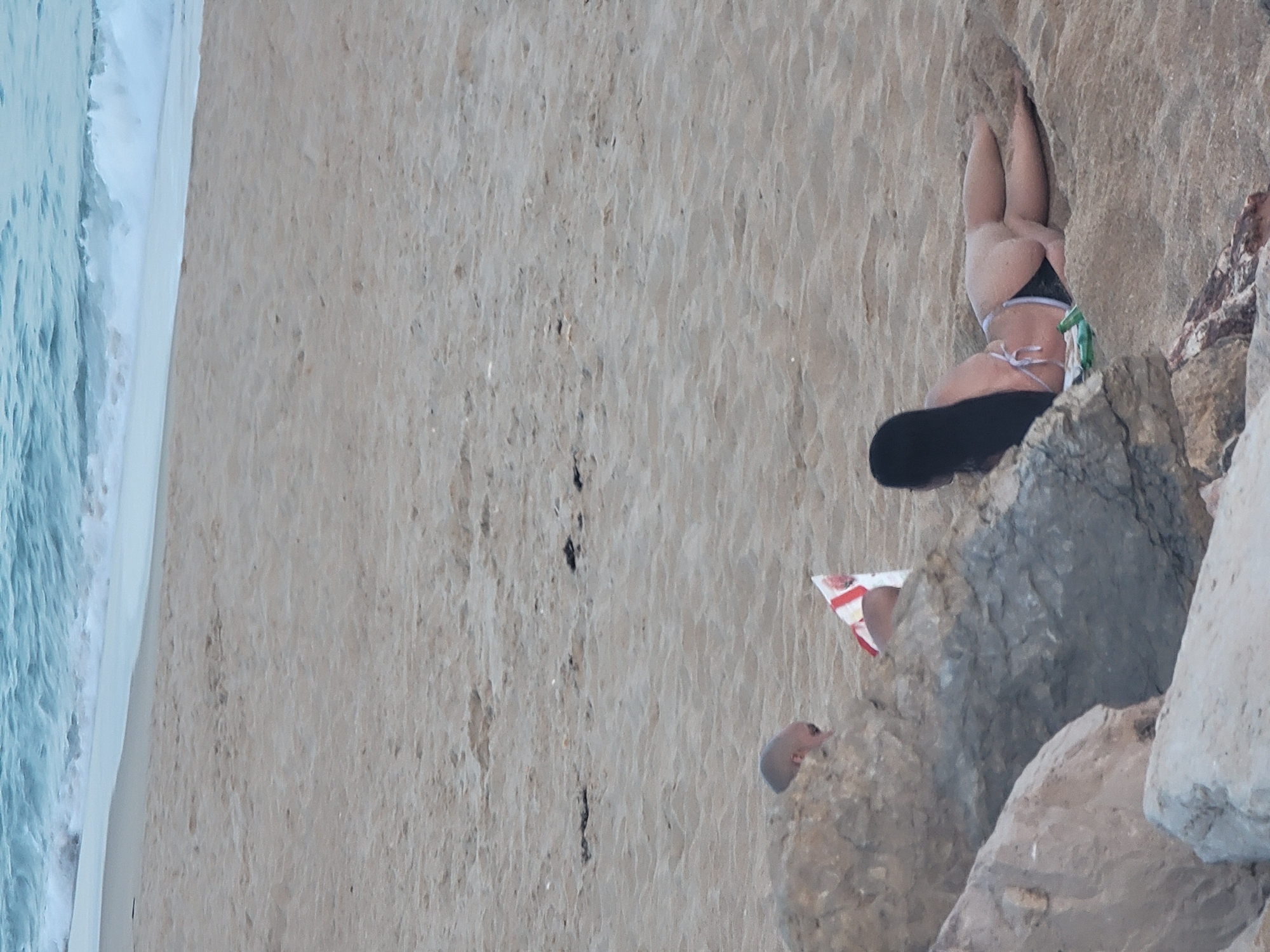 The image depicts a beach scene in Quarteira, Portugal. In the foreground, large rocks are visible. In the middle ground, a person with a shaved head is partially obscured by the rocks, facing away from the camera. Next to them is a red and white object. To the right of the rocks, a person with long dark hair is lying on their stomach on the sand, wearing a bikini. Their legs are extended and positioned on the sand. A green bottle is lying on the sand near the person. In the background, the ocean with breaking waves and a sandy shore are visible.
