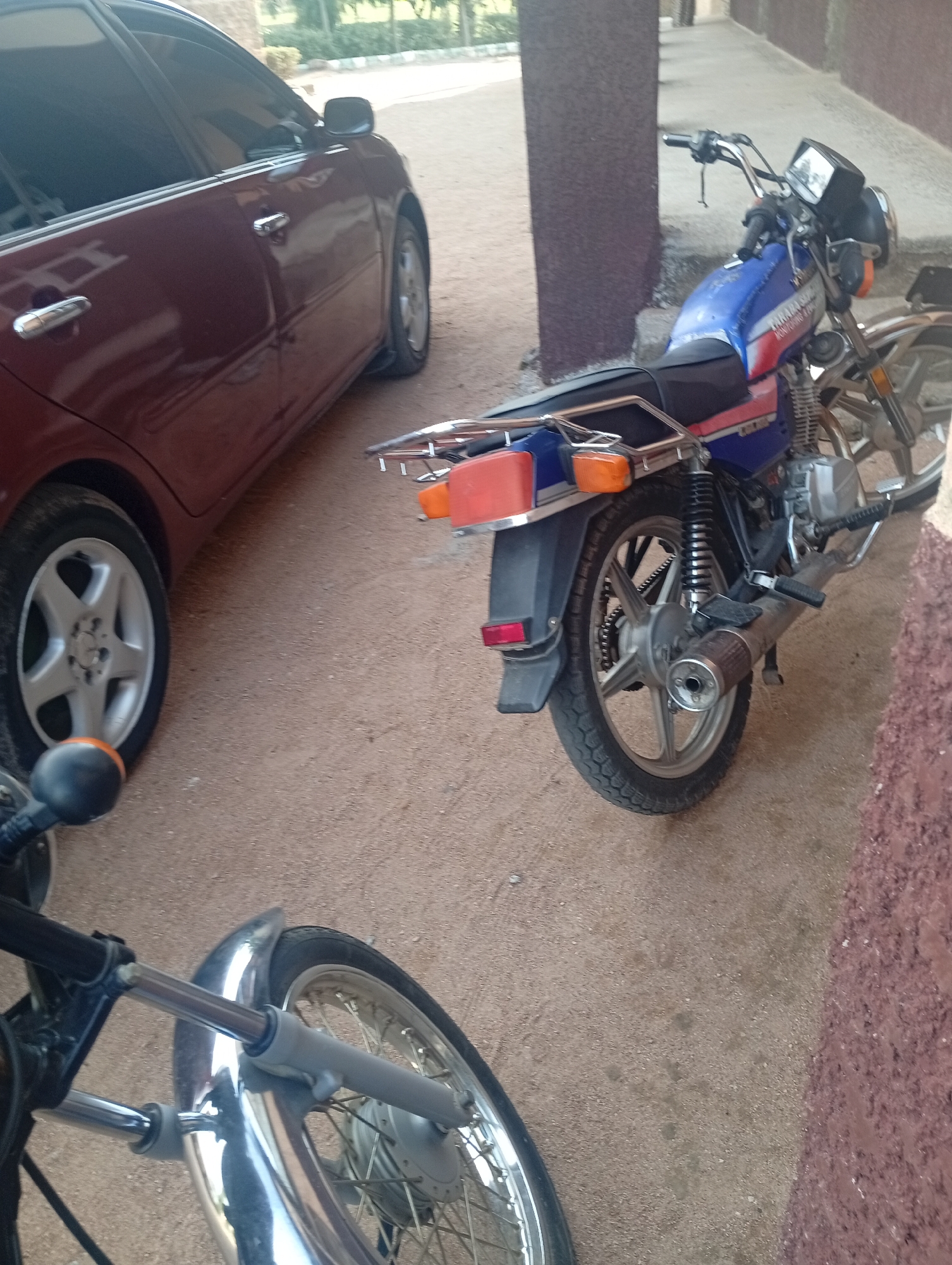 Two motorcycles and a maroon sedan are parked on a sandy, outdoor surface in Jalingo, Nigeria. The motorcycle in the foreground is black with silver accents and its front wheel is visible. The second motorcycle, parked to the right and slightly behind the car, is blue and red. It has a silver exhaust pipe, black seat, and silver wheels. The car is a sedan with tinted windows and silver rims. The scene appears to be in a courtyard or open area with a building with brown columns and a walkway in the background. The lighting suggests it is daytime. There is no visible text in the image.