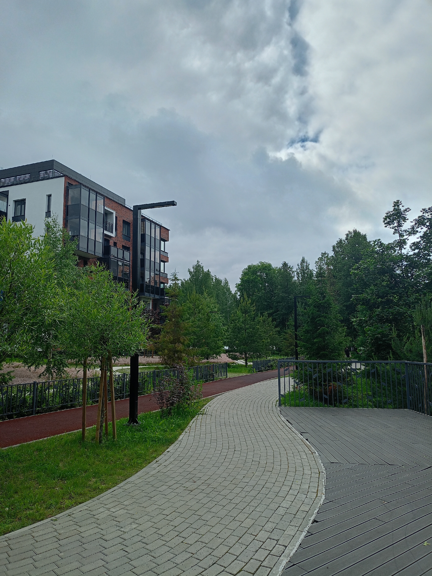 The image depicts an outdoor scene in what appears to be a modern urban development. On the left, a multi-story building with white and brick facades and large glass windows is visible. In front of the building, there is a paved walkway with grey interlocking bricks that curves to the right. Adjacent to the walkway is a red running track. Several trees with green foliage are planted along the walkway and running track. A dark grey metal fence runs along the right side of the image, separating the walkway from a wooden deck. The deck is also grey and features a geometric pattern. Beyond the fence and deck, there is a dense stand of green trees. The sky is overcast with a mix of grey and white clouds. The scene is static, with no people or animals present. The environment suggests a park-like setting integrated with residential or commercial buildings.