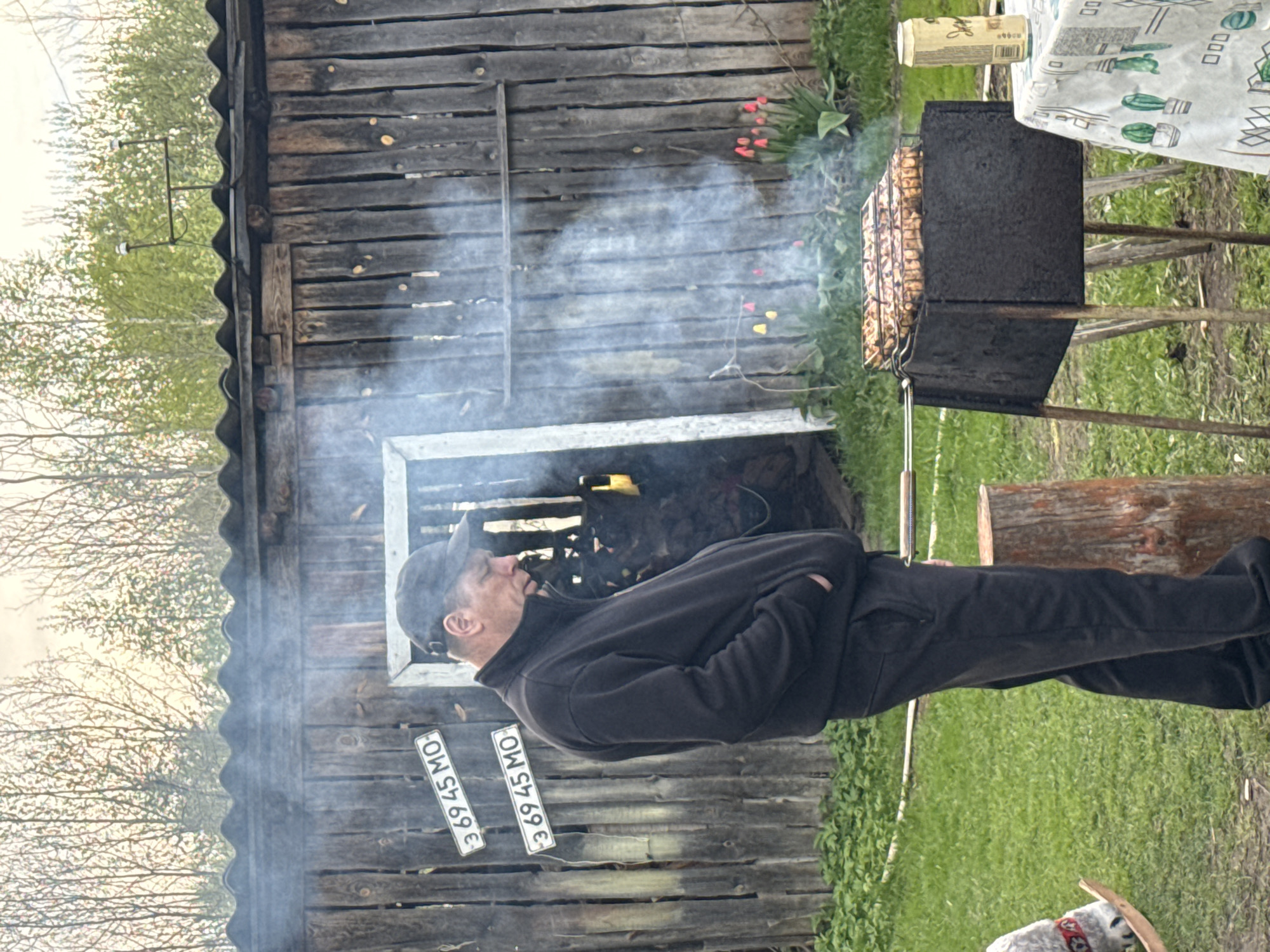 A middle-aged man wearing a black zip-up jacket and a dark baseball cap stands with his hands in his pockets, watching a barbecue grill. The grill is filled with meat skewers and is located near a weathered wooden shed or barn-like structure. The shed has two car license plates with "36945 MO" attached to it. There is smoke emanating from the grill, obscuring parts of the background. The setting is a grassy outdoor area. This scene is potentially in Красный Маяк, Russia.