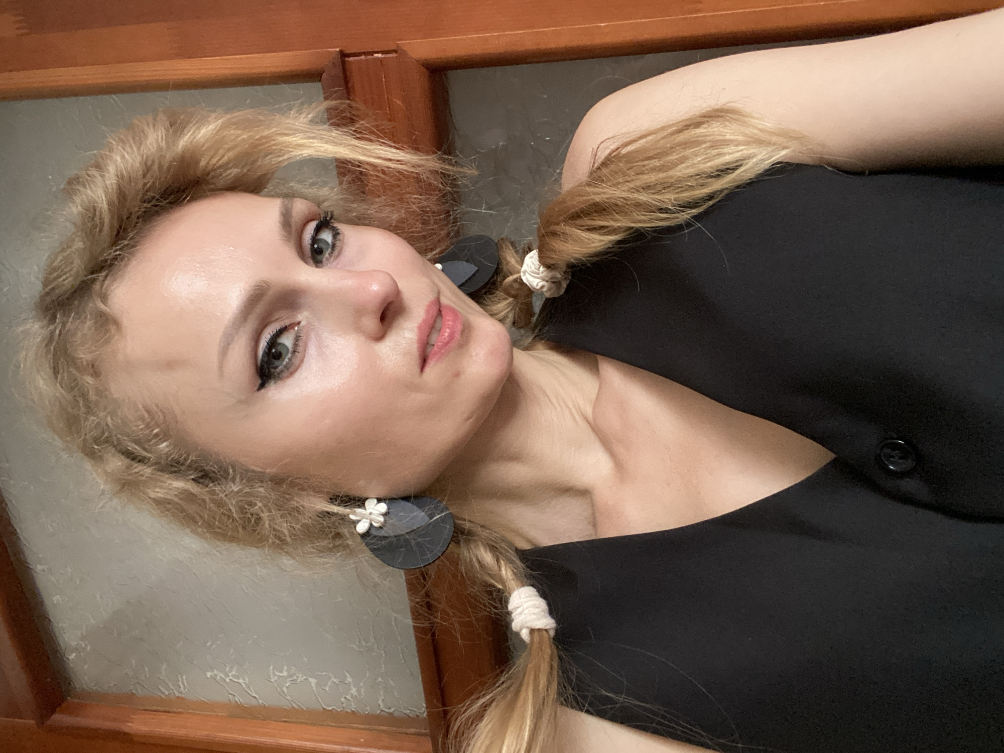 A blonde woman with braided pigtails and large circular earrings with floral accents is looking at the camera. She is wearing a black v-neck vest with a single button. The background appears to be an interior with a textured wall and a wooden door frame. The image was taken in Lida, Belarus.