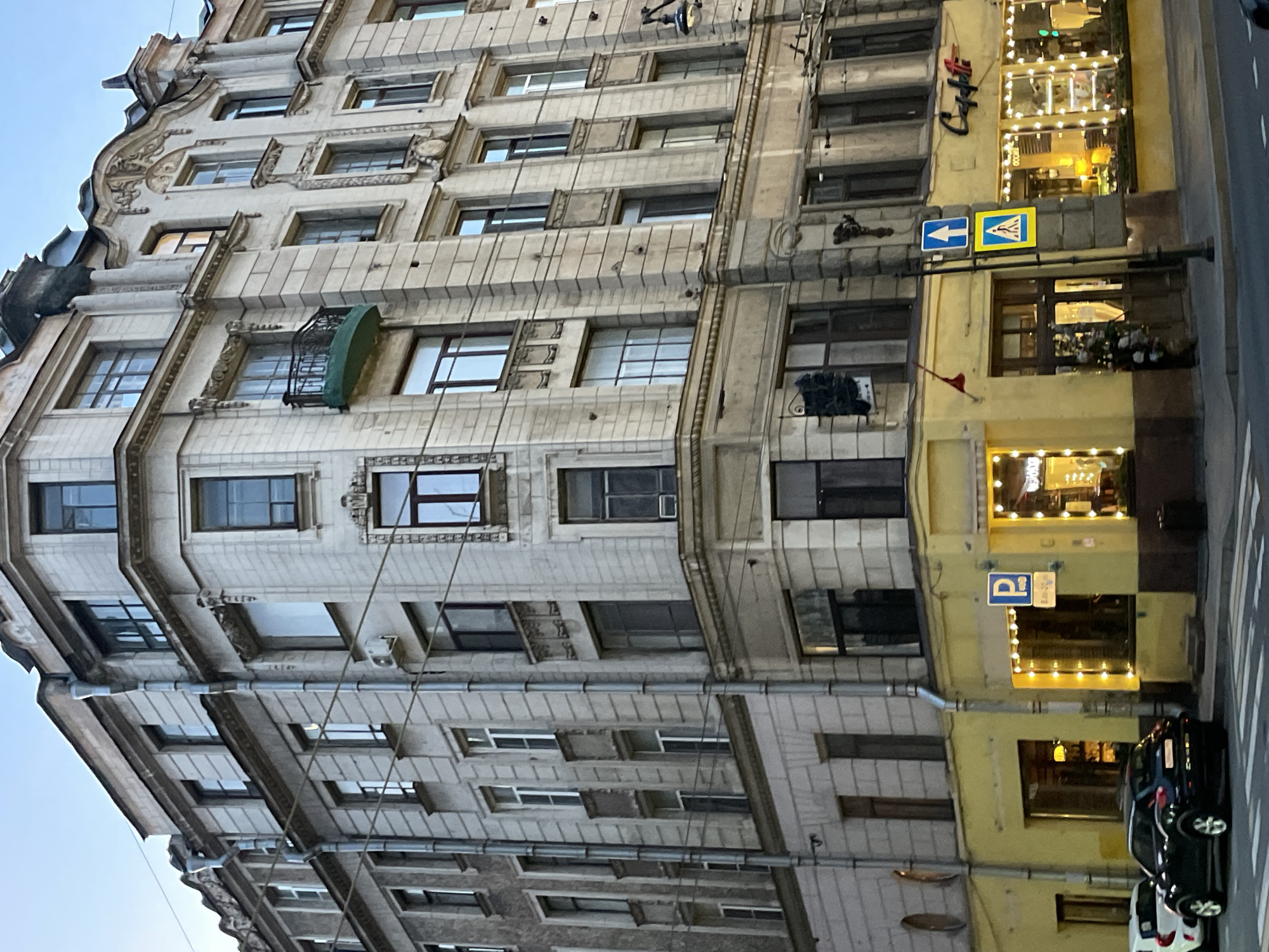 The image depicts a multi-story building facade in Saint Petersburg, Russia. The building features ornate architectural details, including decorative moldings and window frames. To the right, a yellow-painted section houses a café with illuminated signs. Street signs for pedestrians and parking are also visible. Several cars are parked along the street. The overall environment suggests an urban setting, likely within the historic center of Saint Petersburg.
