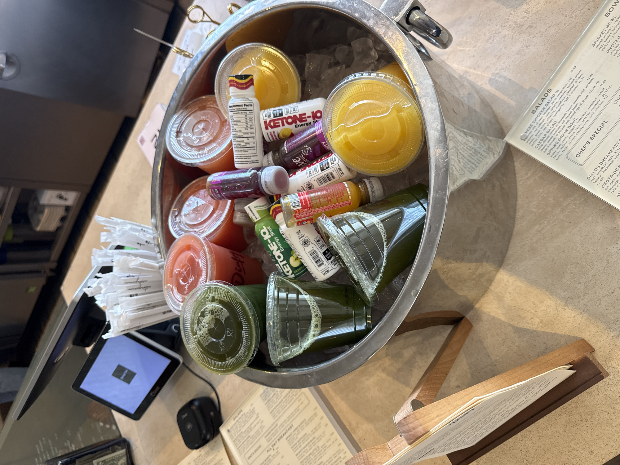Here is a description of the image:
**Scene**: The image shows a tabletop display of beverages and food options, likely at a restaurant or cafe.
**Objects**:
* A metal ice bucket containing an assortment of drinks, including juices and energy shots. Notable brands include Ketone-IQ. The drinks are surrounded by ice.
* A payment terminal (likely a tablet) showing a display with a blue background.
* A stack of wrapped straws.
* A partially visible menu with items listed under headings like "SALADS," "BOWLS," and "CHEF'S SPECIAL." Specific items like "NESE CHICKEN SALAD," "BRISKET BOWL," and "DIALOG BREAKFAST" are readable.
* A wooden menu holder.
**Environment**: The setting is indoors, likely a cafe or restaurant. The tabletop surface is a light color, possibly a type of stone or composite.
**Inferences**: The scene suggests a point of sale or service area where customers might select and pay for items. The variety of drinks indicates a focus on health-conscious or energizing options. The menu suggests a modern, casual dining establishment.
**Location**: While the precise location in Los Angeles is difficult to ascertain from the image alone, the menu item names and food/beverage offerings suggest a trendy or upscale area of the city.
