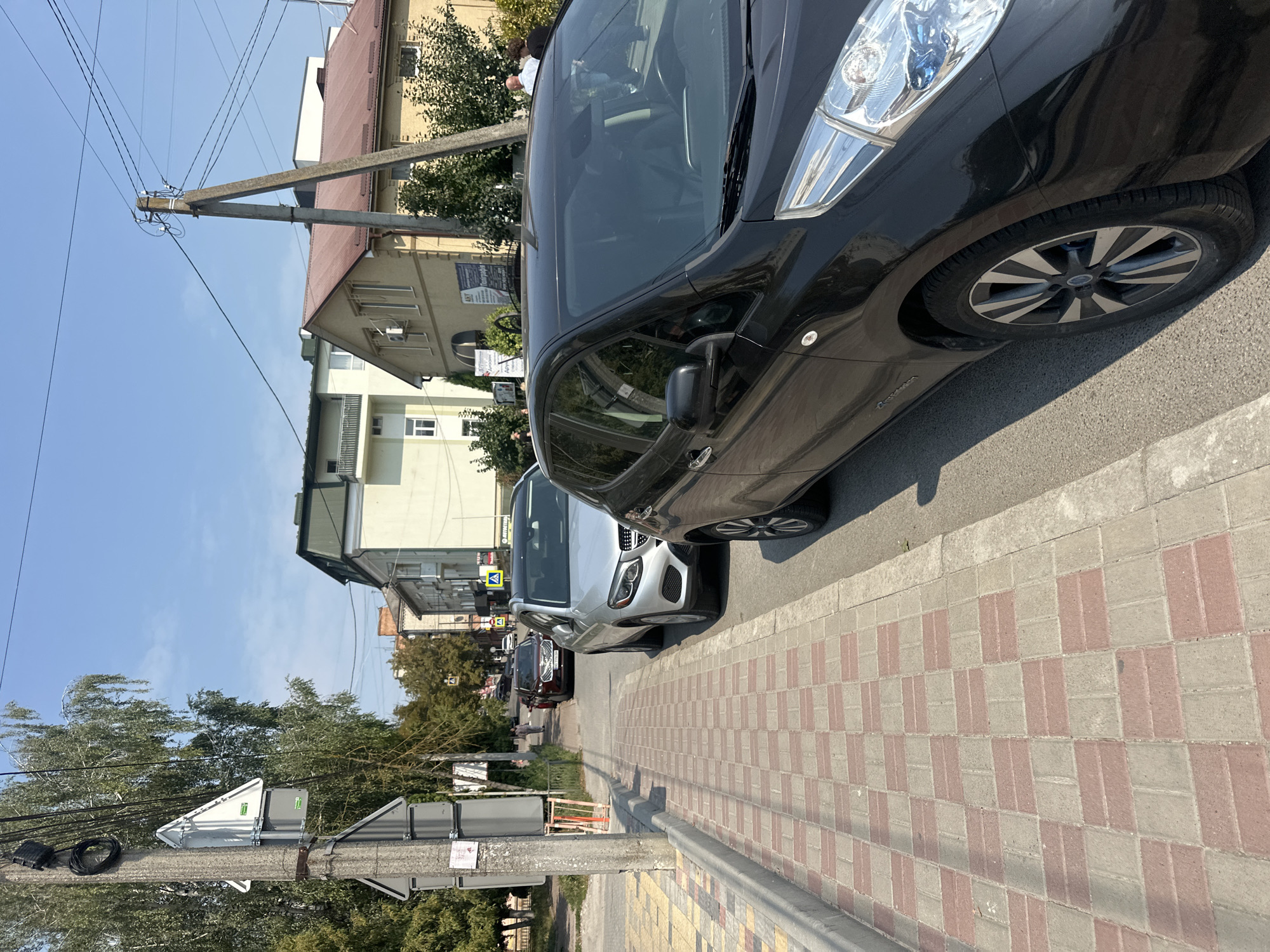The image depicts a street scene in Ternopil, Ukraine, on a sunny day. In the foreground, a black electric car is parked on the right side of the road. Adjacent to the car is a sidewalk paved with red and gray rectangular tiles. The sidewalk is bordered by a curb and the road.
Behind the black car, a silver Mercedes-Benz SUV is parked. Further down the street, a dark red car is also visible. A few pedestrians are walking on the sidewalk in the background.
To the left of the sidewalk, a utility pole is prominent, with various wires and attachments. Two triangular signs are mounted on the pole, one above the other. Trees line the left side of the street.
In the background, residential and commercial buildings are visible, with some architectural details such as balconies and windows. The sky is clear and blue, indicating fair weather. The overall impression is a typical urban street scene in Ukraine.