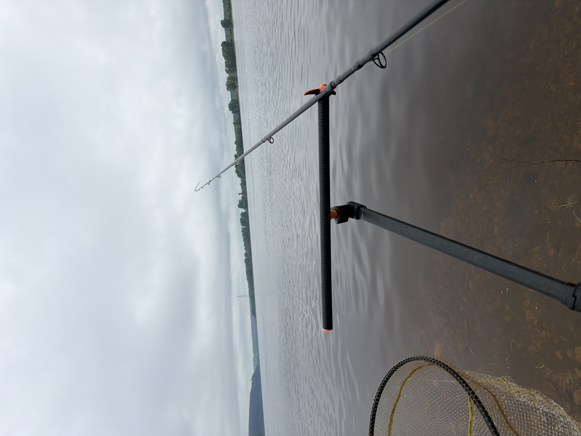 This image captures a serene fishing scene on a wide body of water, likely a river or large lake, under an overcast sky. The perspective is from the shore, looking out across the water.
The primary subject is a dark-colored fishing rod, angled across the frame from the lower right to the upper left. It rests on a black rod holder with an orange accent. The rod features multiple eyelets, through which a light-colored fishing line is threaded. The handle, wrapped in a black, ribbed grip, is prominent in the foreground.
Partially visible in the shallow water near the bank is a fishing net with a black frame and yellow mesh, ready for use. The water in the immediate foreground is clear enough to reveal a rocky or pebbly bottom, suggesting a natural shoreline.
Beyond the rod, the expanse of water shows subtle ripples on its surface. A distant shoreline, densely populated with green trees, stretches horizontally across the horizon. Above the tree line, the sky is a uniform, light gray, indicating heavily overcast weather. A faint, distant structure, possibly a power line tower, can be seen on the left horizon.
The image conveys a quiet, contemplative atmosphere typical of a fishing excursion in Chaikovsky, Russia. The cloudy conditions suggest a cool or mild day. There is no visible text in the image.