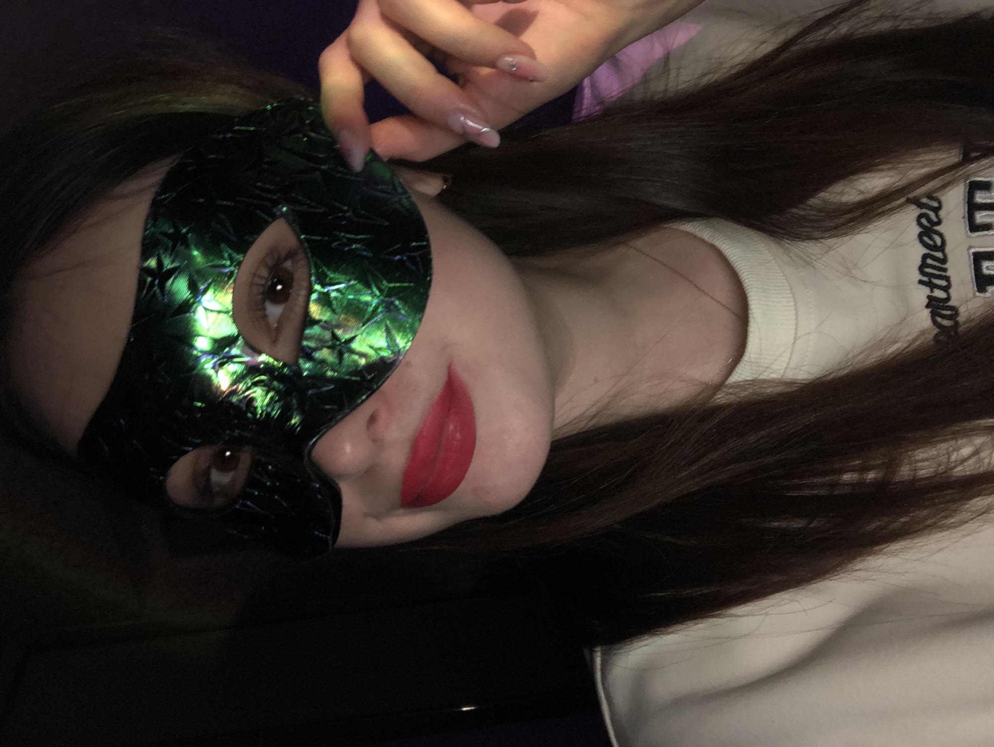 The image displays a young woman in an indoor setting, contextually located in Noviy, Russia. She is captured at an angle, with her head tilted, suggesting a casual or posed moment. The central focus is a vibrant, iridescent green mask with a patterned, possibly star-like texture, which covers the upper half of her face. Her brown eyes are visible through the mask's openings, and she has bright red lipstick on her mouth, forming a slight, pleasant smile. One of her hands, featuring long, manicured nails with a light polish and subtle artistic details, is visible near her temple, seemingly adjusting or holding the mask. Her long, dark brown hair falls over her shoulders and chest. She is wearing a white crew-neck sweater or sweatshirt, with the partial word "artment" embroidered in dark thread on the front, likely part of "Apartment" or "Department." The background is dark and indistinct, suggesting dim lighting, possibly at night or in a low-light environment, with a faint hint of purple or pink on the left side. The overall mood is playful or mysterious.