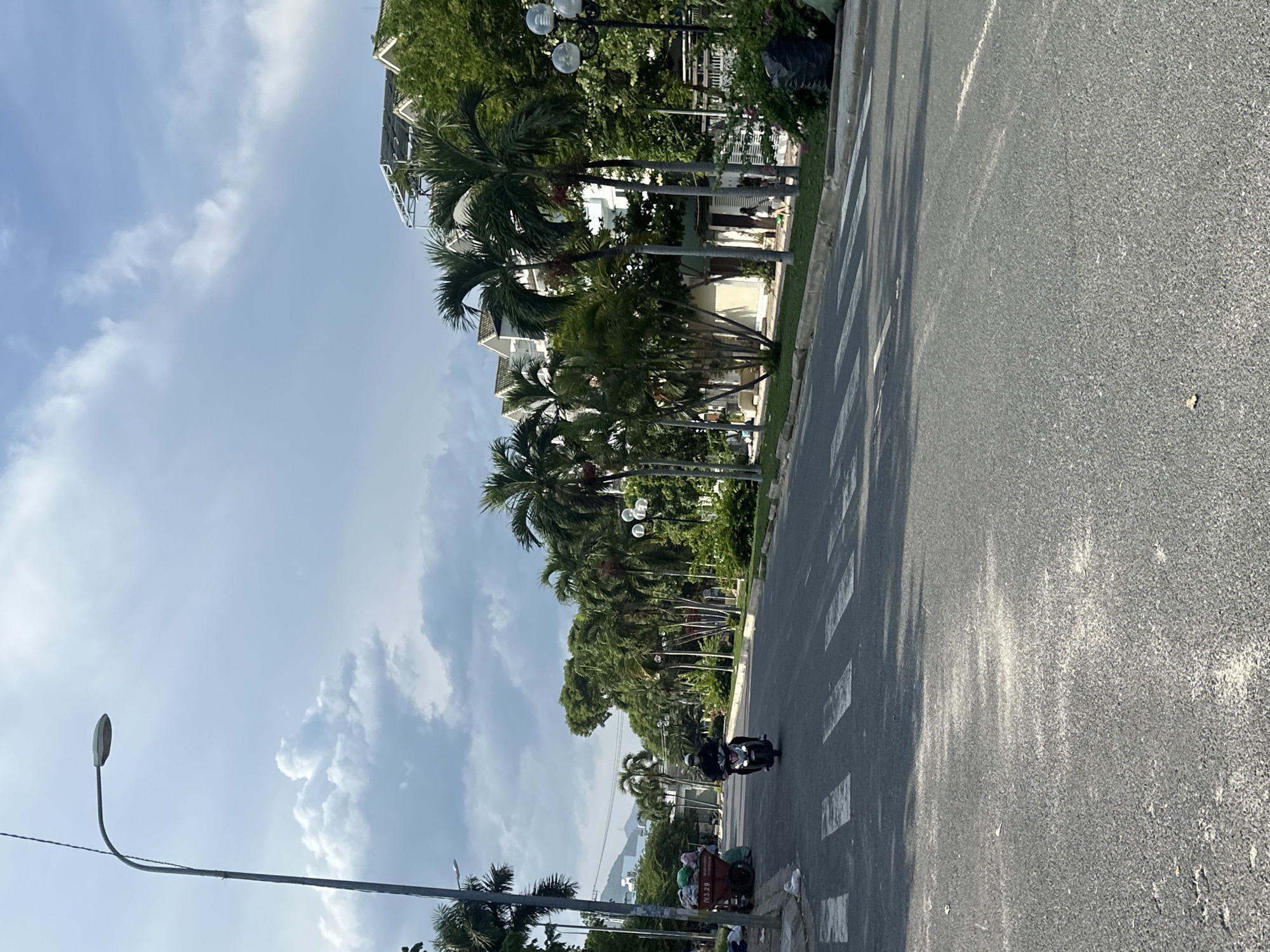 The image depicts a street scene in Nha Trang, Vietnam, likely during the daytime. A paved road with white pedestrian crossing markings runs through the center of the frame, curving to the right. On the left side of the road, a person rides a scooter. A red refuse cart filled with various items is parked on the left shoulder, adjacent to some white plastic bags. A tall streetlamp stands on the left side of the road. The right side of the road is lined with palm trees and other greenery, interspersed with multi-story buildings that appear to be residences or hotels. The sky is partially cloudy.