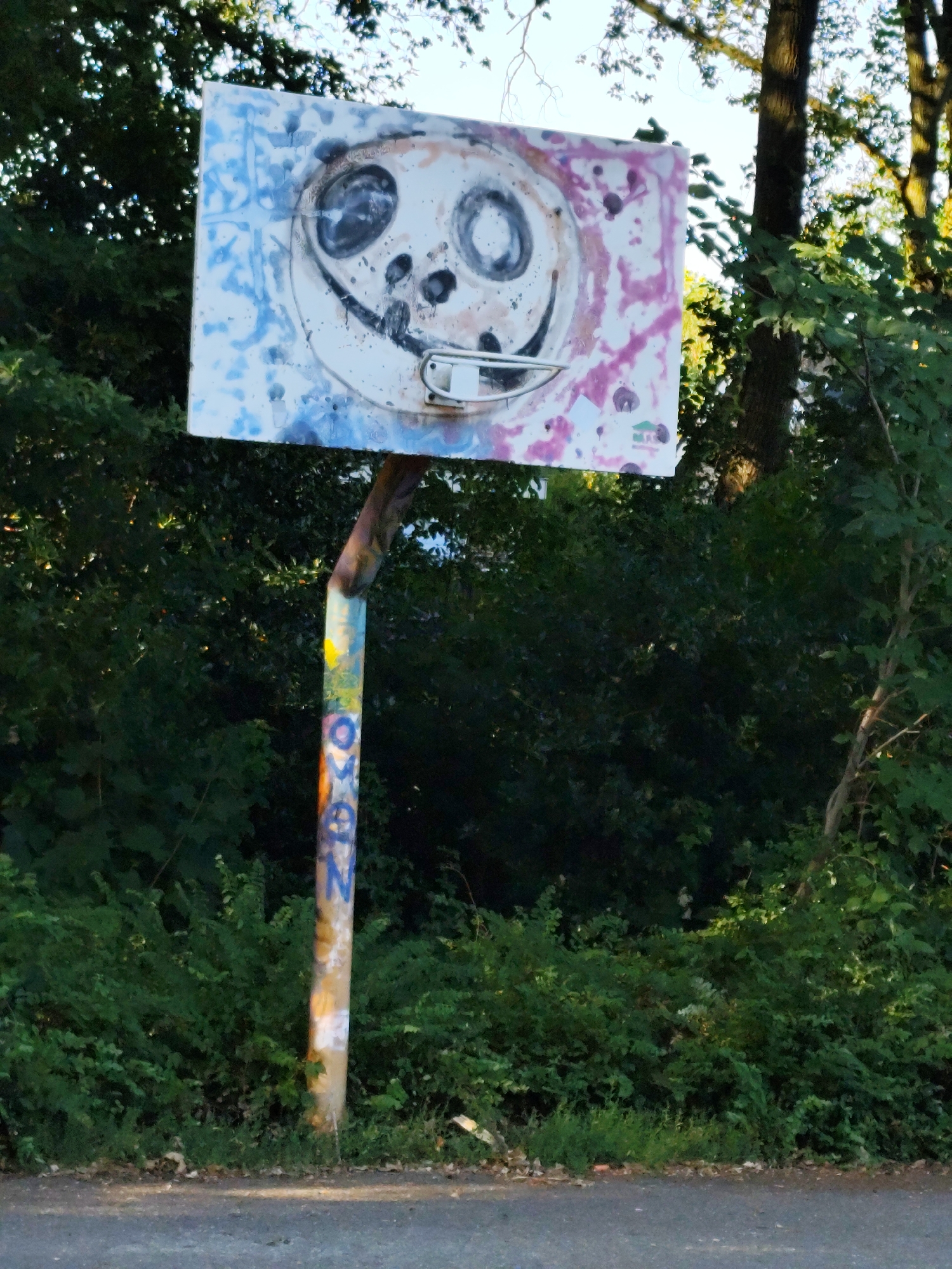 The image shows a weathered basketball backboard with a painted smiley face, located in a natural setting with lush green trees and bushes. The backboard itself is rectangular with rounded corners and features a stylized, somewhat abstract depiction of a face with large, circular eyes and a wide smile. Splashes of blue and pink paint are visible on the backboard, suggesting it has been vandalized or artistically altered.
The basketball hoop, a white metal rim, is attached to the backboard. The backboard is supported by a thick, rough-textured pole that is also covered in graffiti. The graffiti on the pole includes letters and symbols in various colors, including orange, yellow, blue, and black. The pole appears to be leaning slightly, indicating potential disrepair.
The background consists of dense foliage, with dark green leaves and branches of trees creating a backdrop. Sunlight filters through the trees, casting dappled light and shadows on the scene. The ground in front of the backboard is a mix of dry leaves and some green grass, with a paved surface, likely a road or pathway, visible in the foreground. The overall impression is one of an abandoned or neglected recreational area integrated into a natural environment.
This basketball backboard is located at the former St. Antonius Church in Schaijk, Netherlands, a site that is now known as 't Kerkpad.