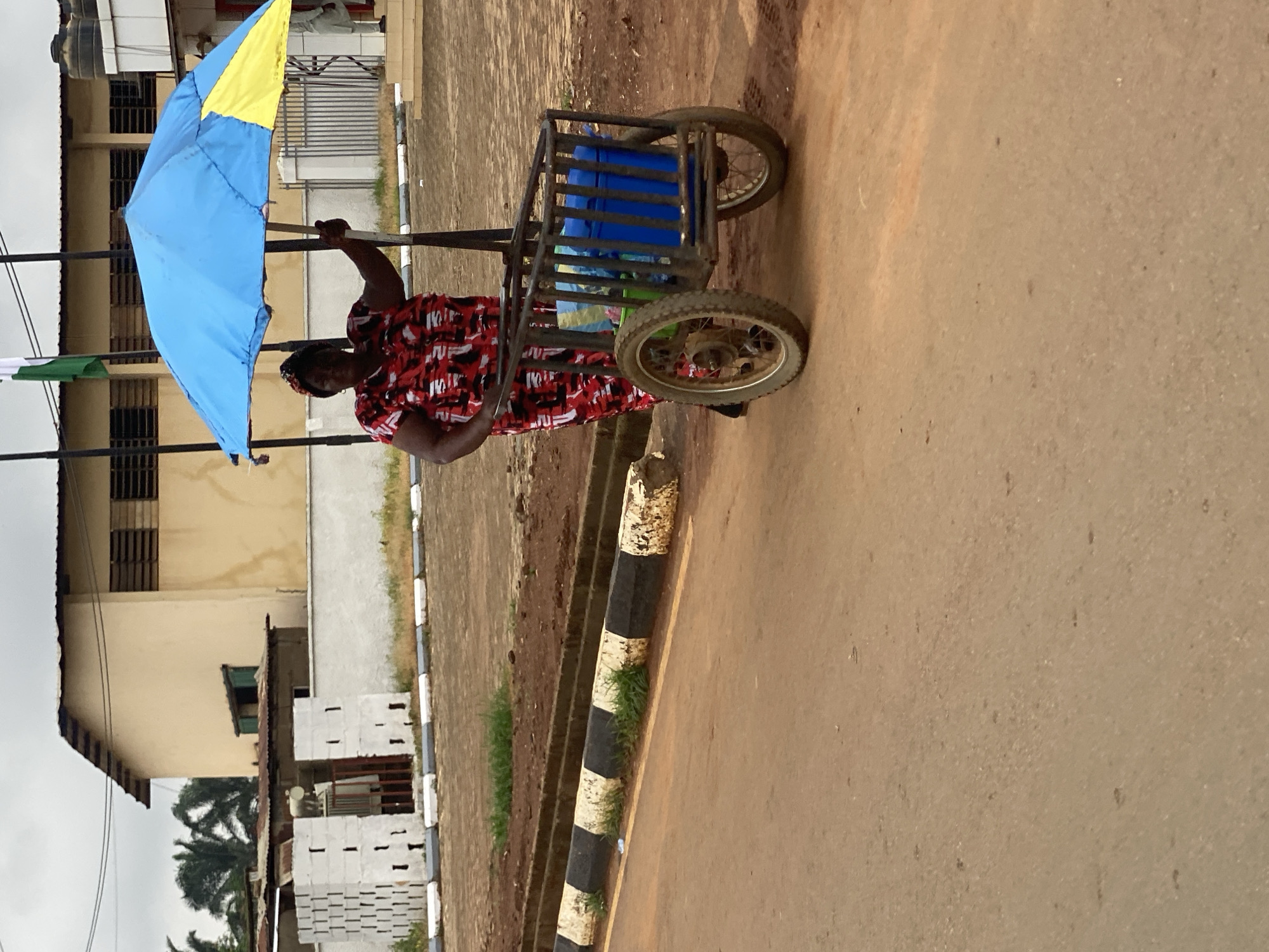 A woman is walking on a paved road in Kafanchan B, Nigeria. She is pulling a two-wheeled cart constructed from a metal frame, which holds blue containers. She is holding a blue and yellow umbrella over her head for shade. She is wearing a red and black patterned dress. The road is next to a curb marked with black and white stripes. A building is behind her, constructed of tan-colored materials with a dark roof. Part of a palm tree is visible in the background.
