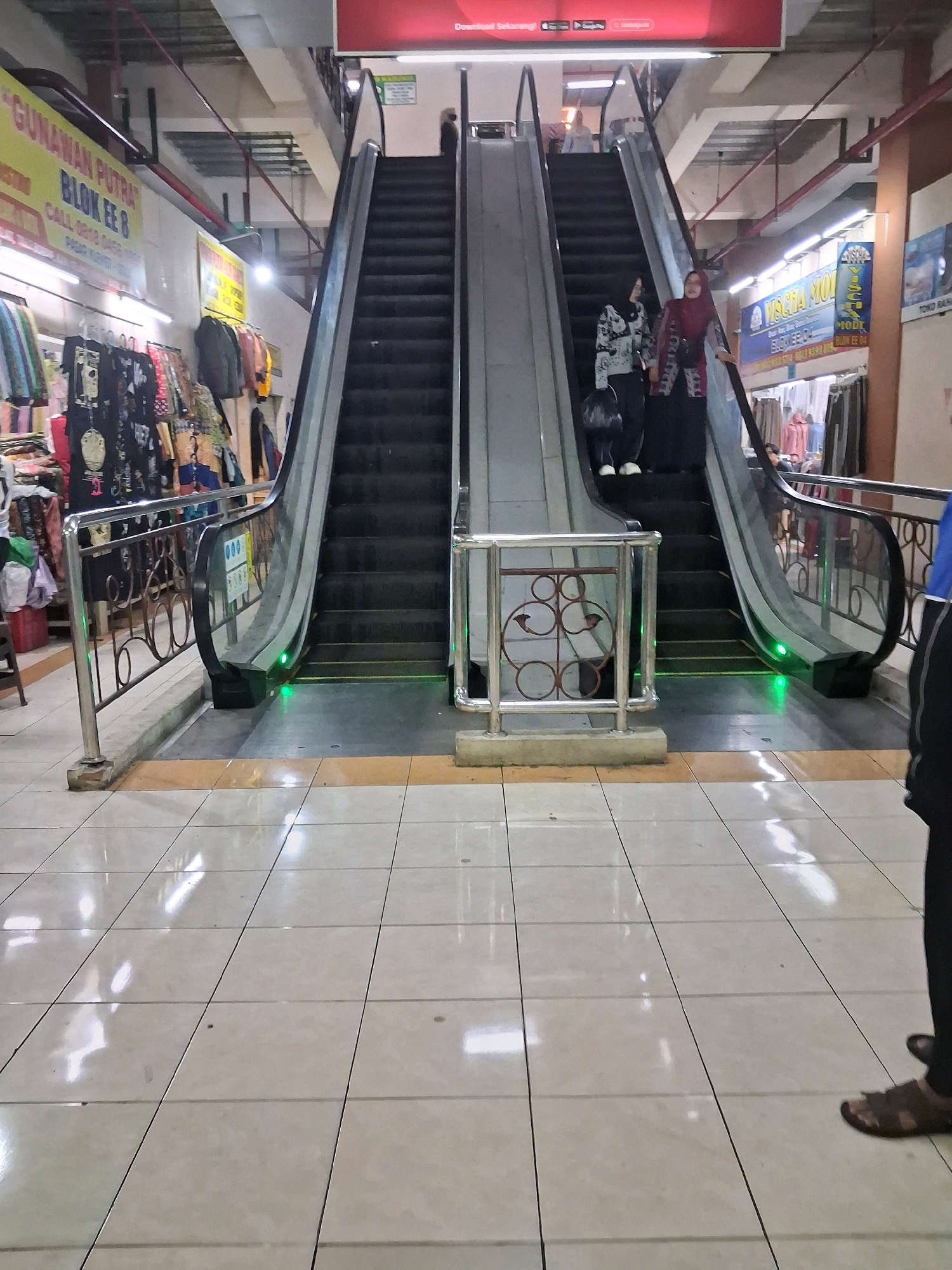The image captures an indoor scene in Surakarta, Indonesia, likely within a shopping center or market, featuring a prominent escalator. Two women are visible on the ascending escalator, one in a maroon hijab and patterned clothing, the other in a patterned top and dark pants. The escalator is flanked by shops, one with clothing racks displaying various garments. The floor is tiled with a light-colored pattern. Signs in Indonesian are visible, including one that reads "GUNAWAN PUTRA BLOK EE 8." Green lights are illuminated at the base of the escalator. Another person, partially visible, is standing on the tiled floor to the right of the image.