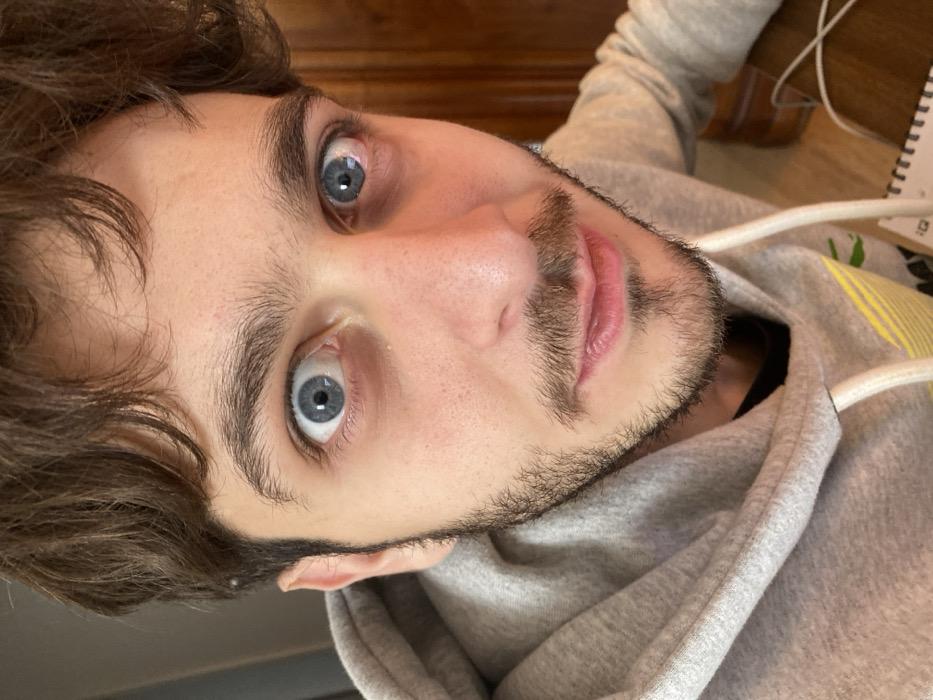 A close-up, head-and-shoulders portrait of a young adult male, reportedly captured in Strasbourg, France. The subject possesses brown, wavy hair, and widely open light blue eyes with noticeable periorbital darkening. A light brown stubble covers his chin, upper lip, and jawline. He is dressed in a light grey hooded sweatshirt featuring a white drawstring and faint yellow-green horizontal stripes on the chest, layered over a dark undergarment. The male looks directly into the camera. The background on the right side of the frame shows a segment of dark brown wood paneling or furniture. On the bottom right, a white spiral-bound notebook and a white cable are partially visible on an underlying surface.