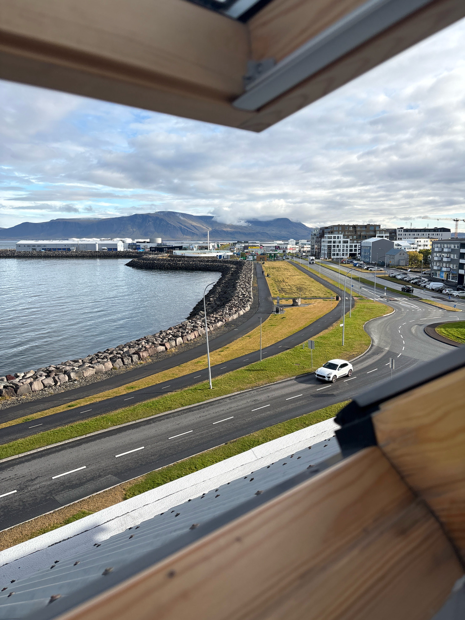 The image shows a coastal view of Reykjavik, Iceland. In the foreground, a wooden architectural element frames the view. Below this, a grey metal roof is visible, with some visible fasteners. Beyond the roof, a paved road curves to the right, with a white car driving on it. The road is bordered by a green grassy strip and a dark grey path, likely a bicycle or pedestrian lane. To the left of the path is a rocky embankment separating it from a body of water.
The water is calm and reflects the cloudy sky. Along the coastline, there are industrial buildings with white facades, and further inland, modern multi-story apartment buildings with dark facades and many windows are visible. In the distance, a mountain range with some cloud cover stretches across the horizon. A construction crane is visible behind the buildings on the right. The sky is overcast with patches of blue visible through the clouds. The overall impression is of an urban coastal environment with a backdrop of natural landscape.