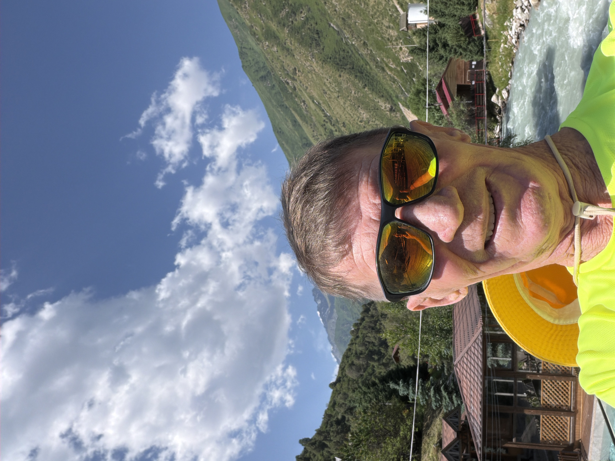 A man wearing sunglasses and a yellow hat takes a selfie outdoors. He is in the foreground, smiling at the camera. Behind him, a river flows through a mountainous landscape. The man is wearing a bright yellow shirt. The sunglasses reflect the surroundings, including part of the man's face and a building. In the background, there are trees, mountains, and several wooden structures, possibly cabins or gazebos. The sky is blue with some white clouds.